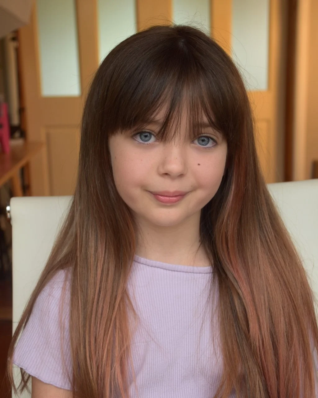 Young girl with long, straight brown hair and blue eyes, wearing a light purple top, sitting in a room with wooden furniture and frosted glass-paneled doors in the background.