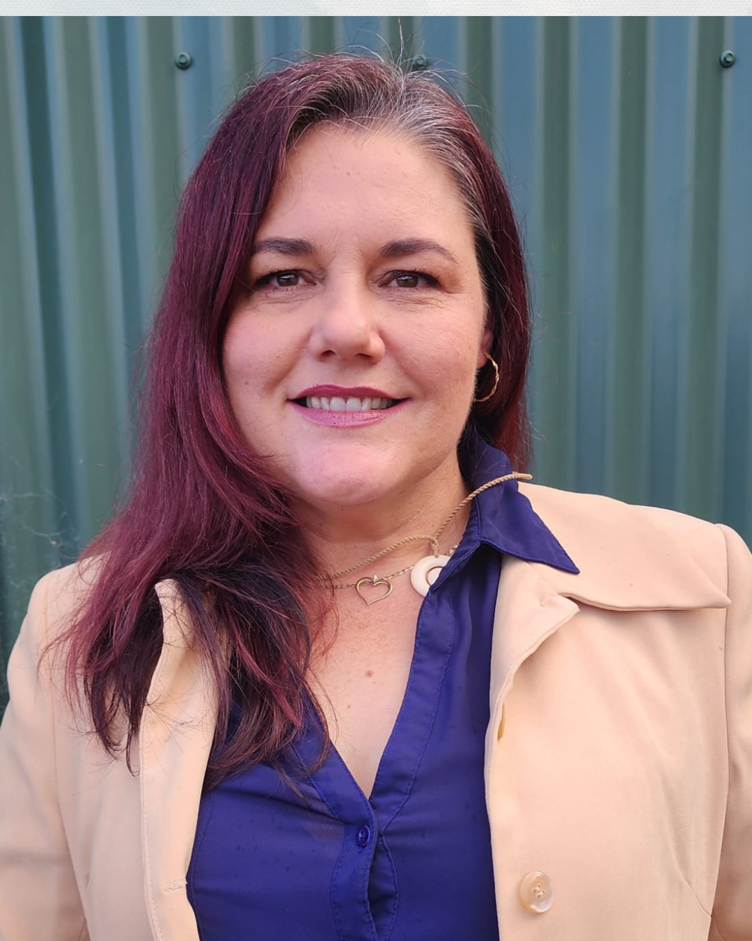 A woman with shoulder-length burgundy hair smiling at the camera in front of a green, corrugated metal background, wearing a beige blazer over a blue shirt, a gold necklace with a heart pendant, and gold hoop earrings.