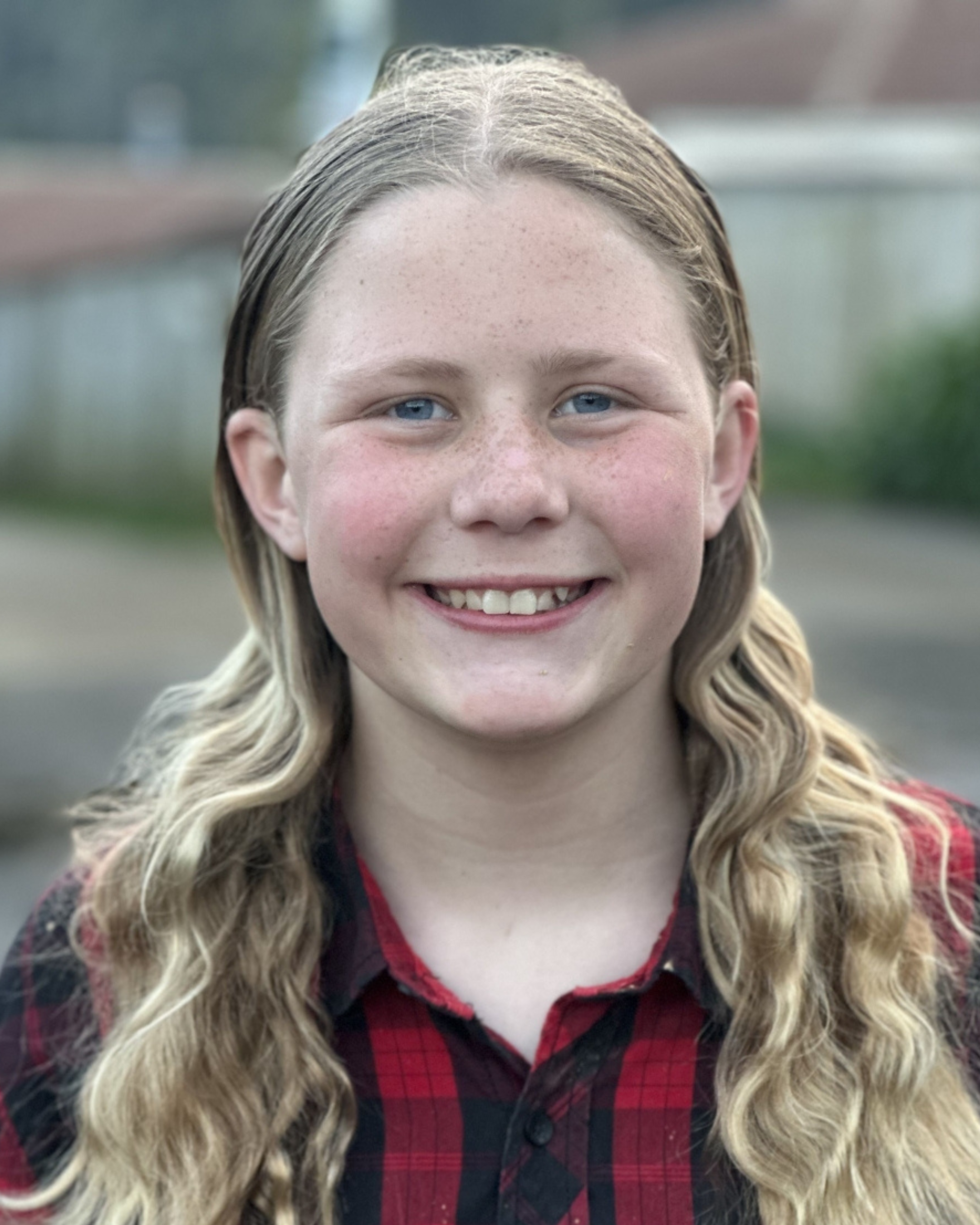 Close-up of a young girl with long, wavy blonde hair, blue eyes, and freckles, smiling outdoors during daytime.