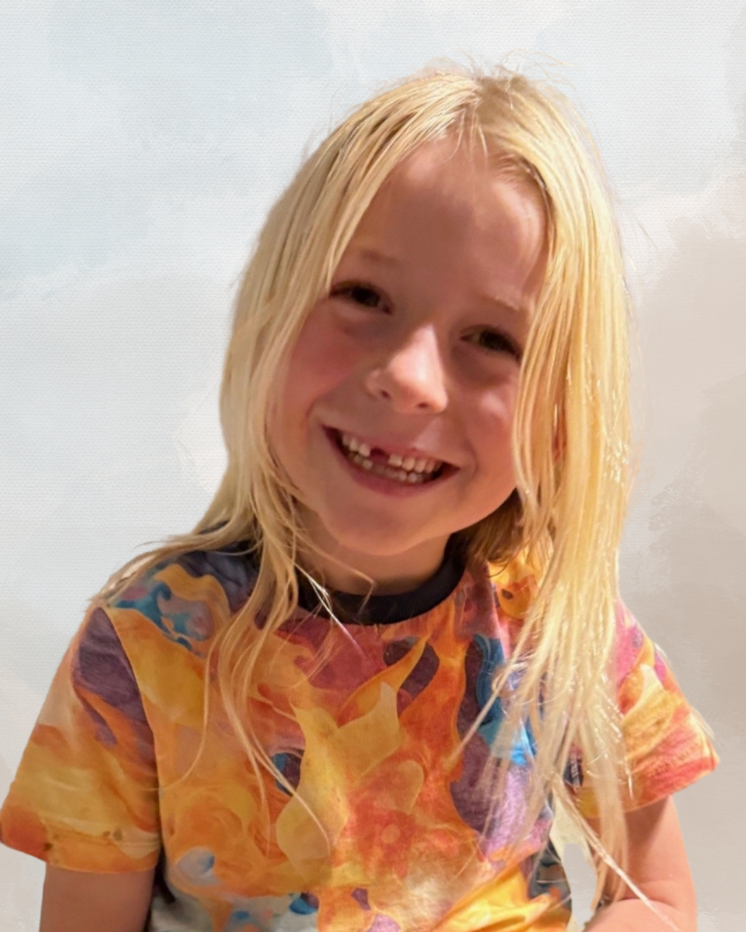 Young girl with long blonde hair smiling, wearing a colorful tie-dye t-shirt, against a pale background.