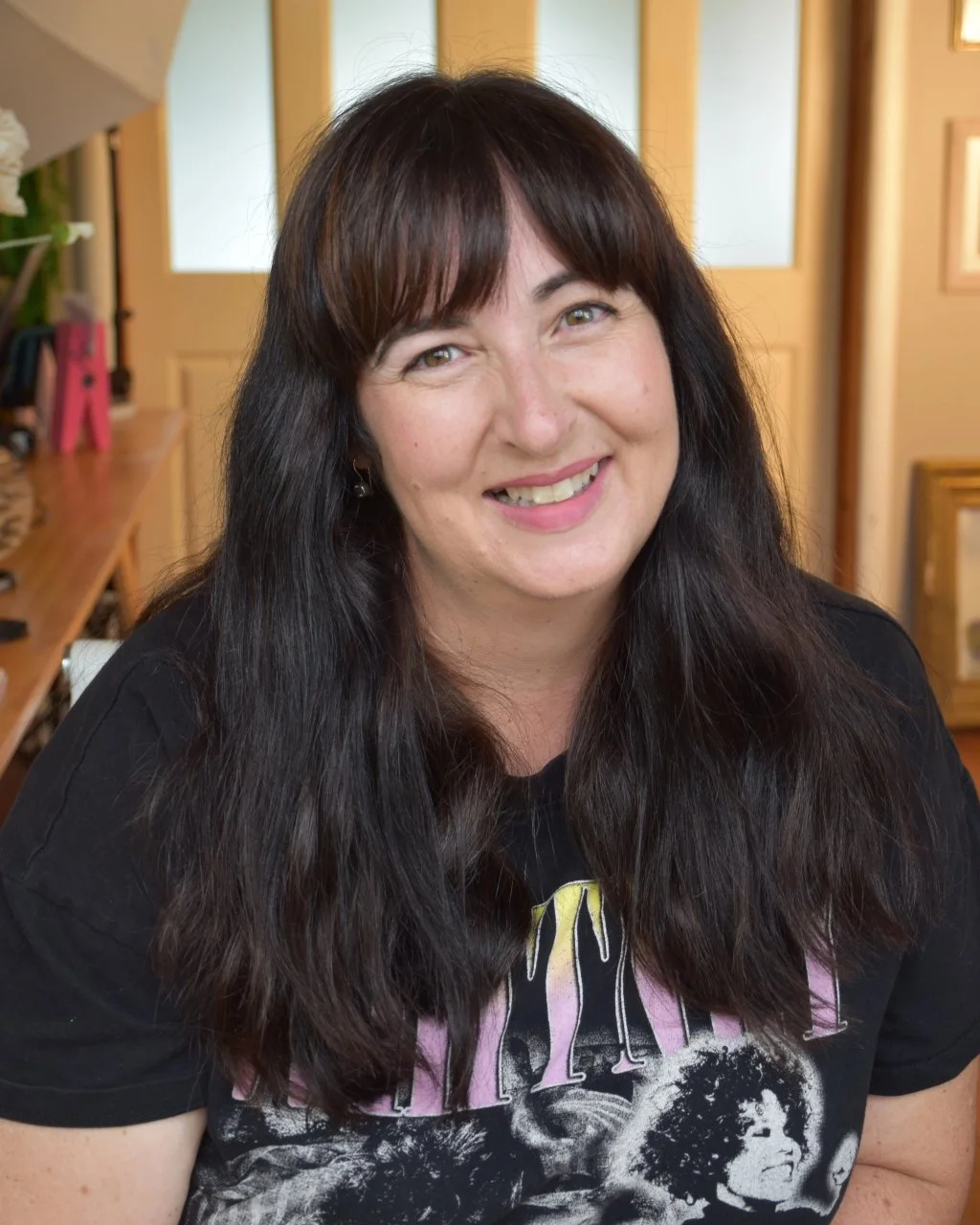A woman with long dark brown hair, smiling, sitting indoors with wooden decor, wearing a black T-shirt with a graphic design.