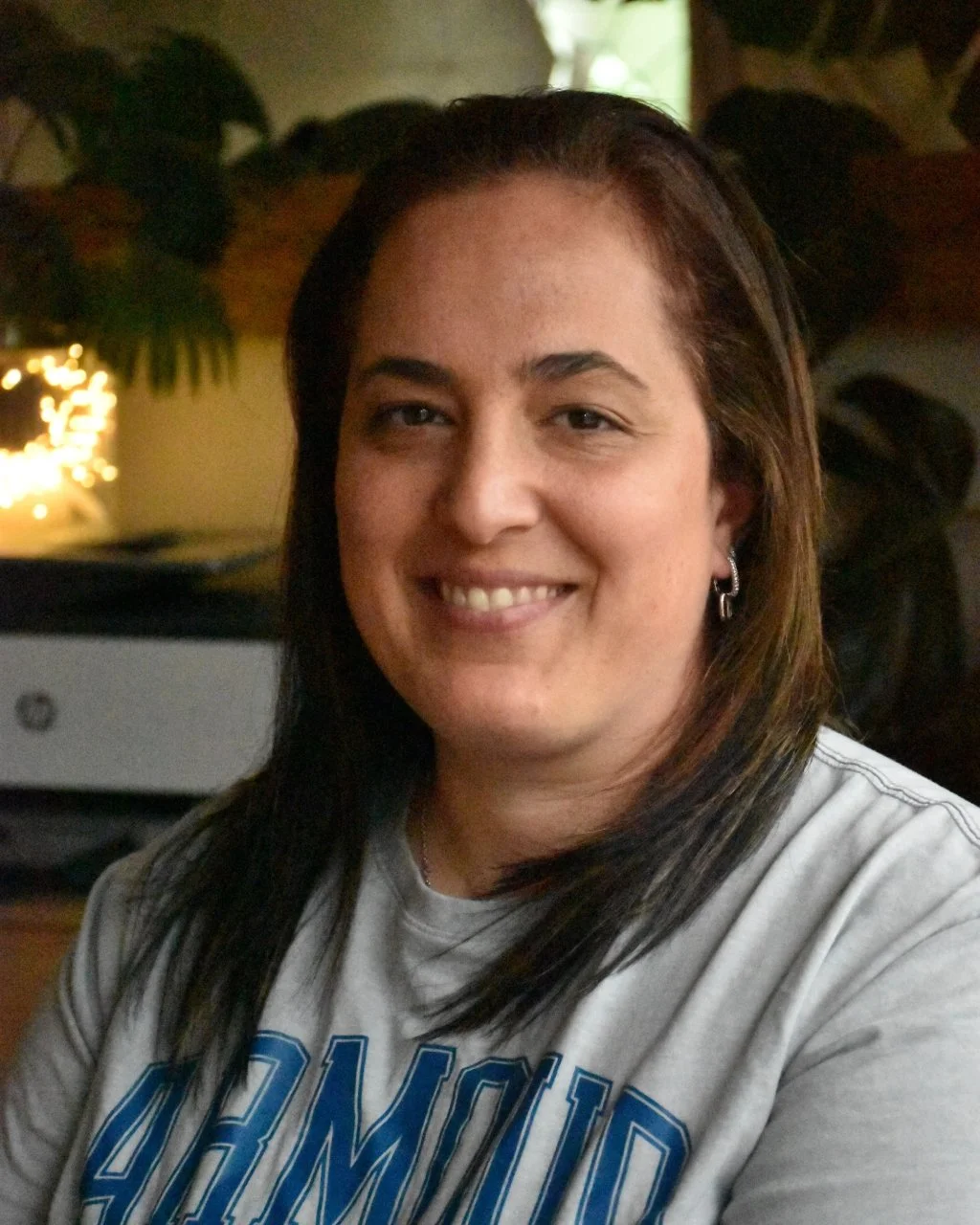 A smiling woman with shoulder-length dark hair with highlights, wearing earrings and a grey T-shirt with blue text, sitting indoors with a blurred background.