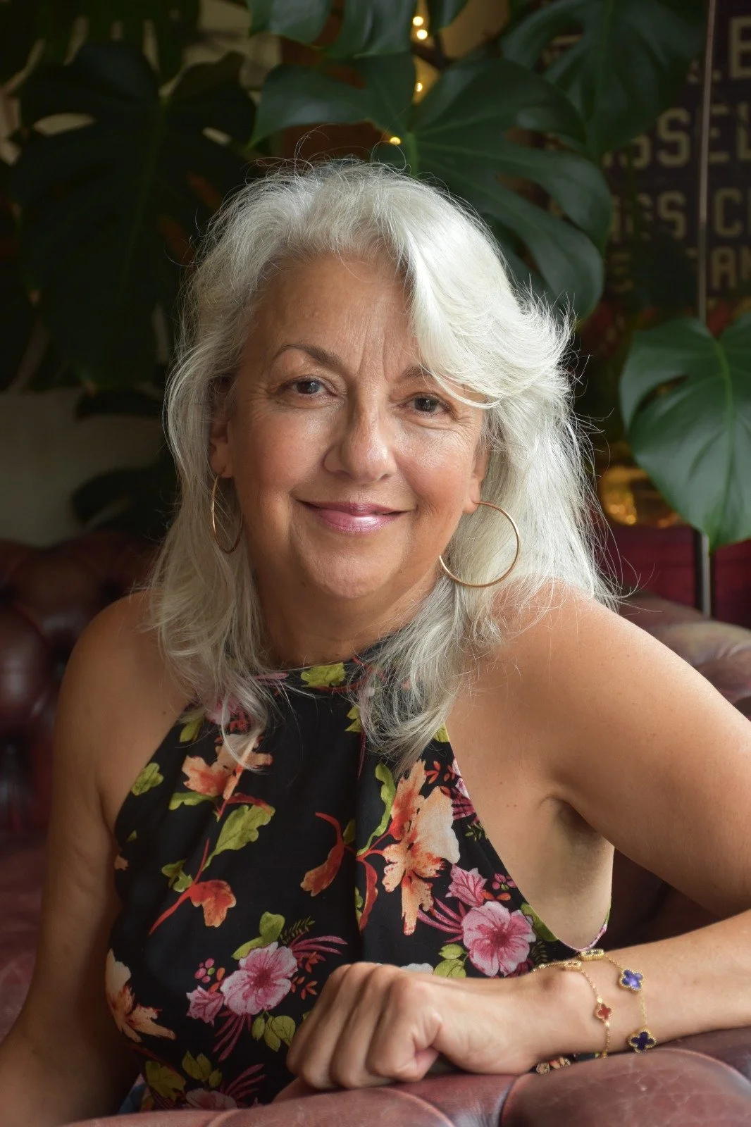 A smiling older woman with shoulder-length gray hair, wearing large hoop earrings and a floral sleeveless dress, sitting on a leather couch with greenery in the background.