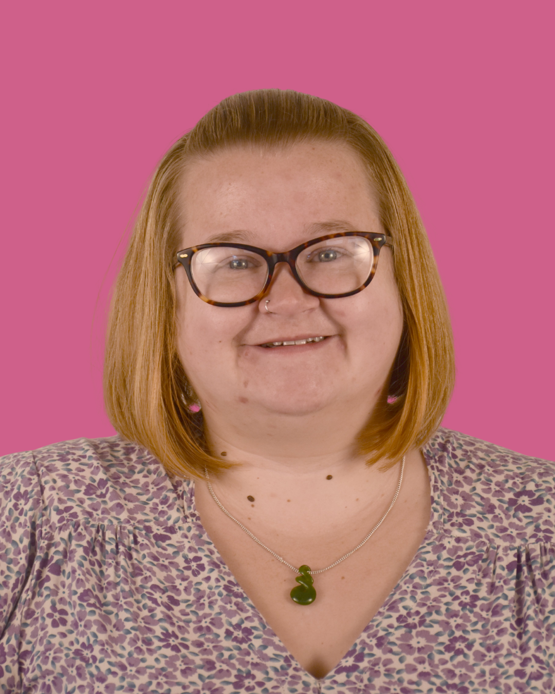 Portrait of a woman with red hair, glasses, and a leopard print top, smiling against a pink background.