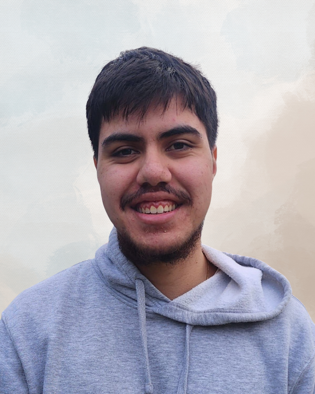 Young man with dark hair, mustache, and beard smiling, wearing a gray hoodie against a cloudy sky background.