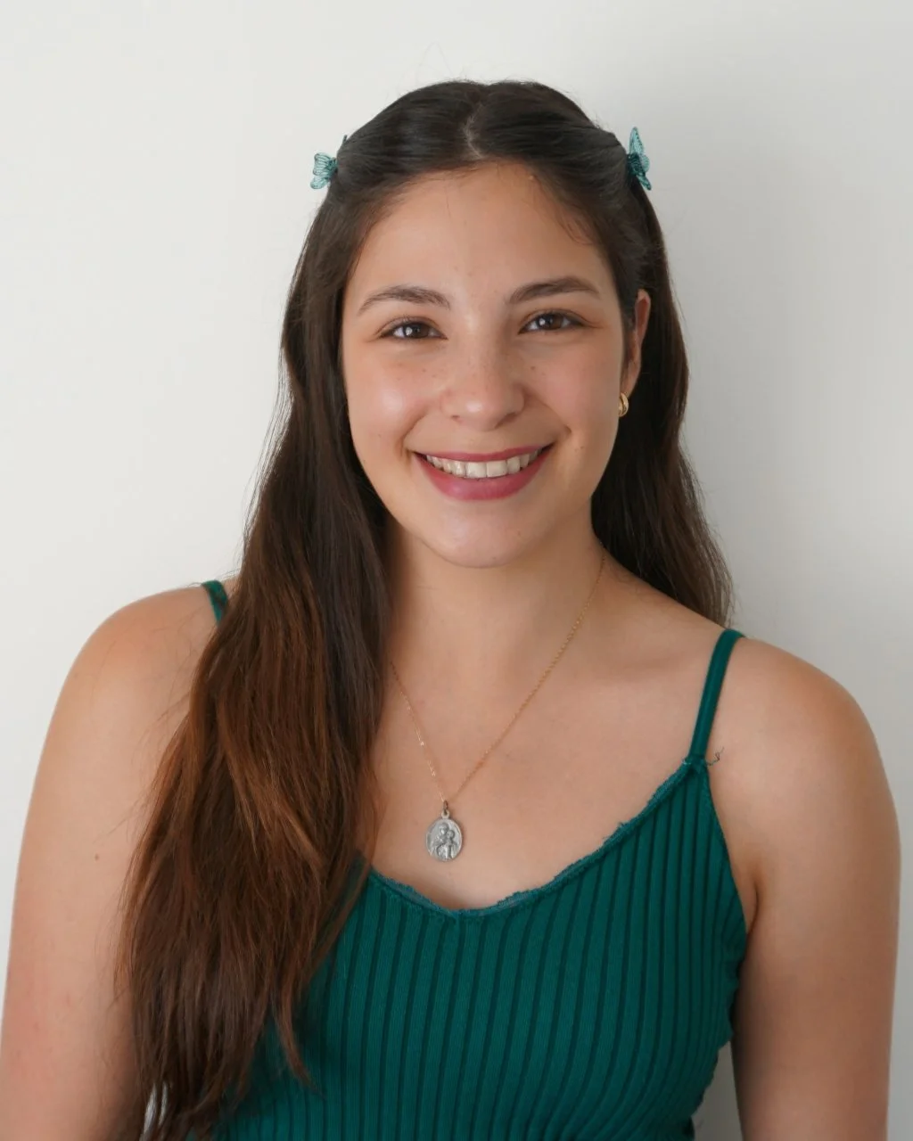 A young woman with brown hair, wearing two teal hair clips, a green striped tank top, and a gold necklace with a religious medal, smiling against a plain white wall.
