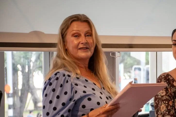 An older woman with blonde hair wearing a light purple dress with black polka dots, holding a pink tablet, standing indoors near a window.