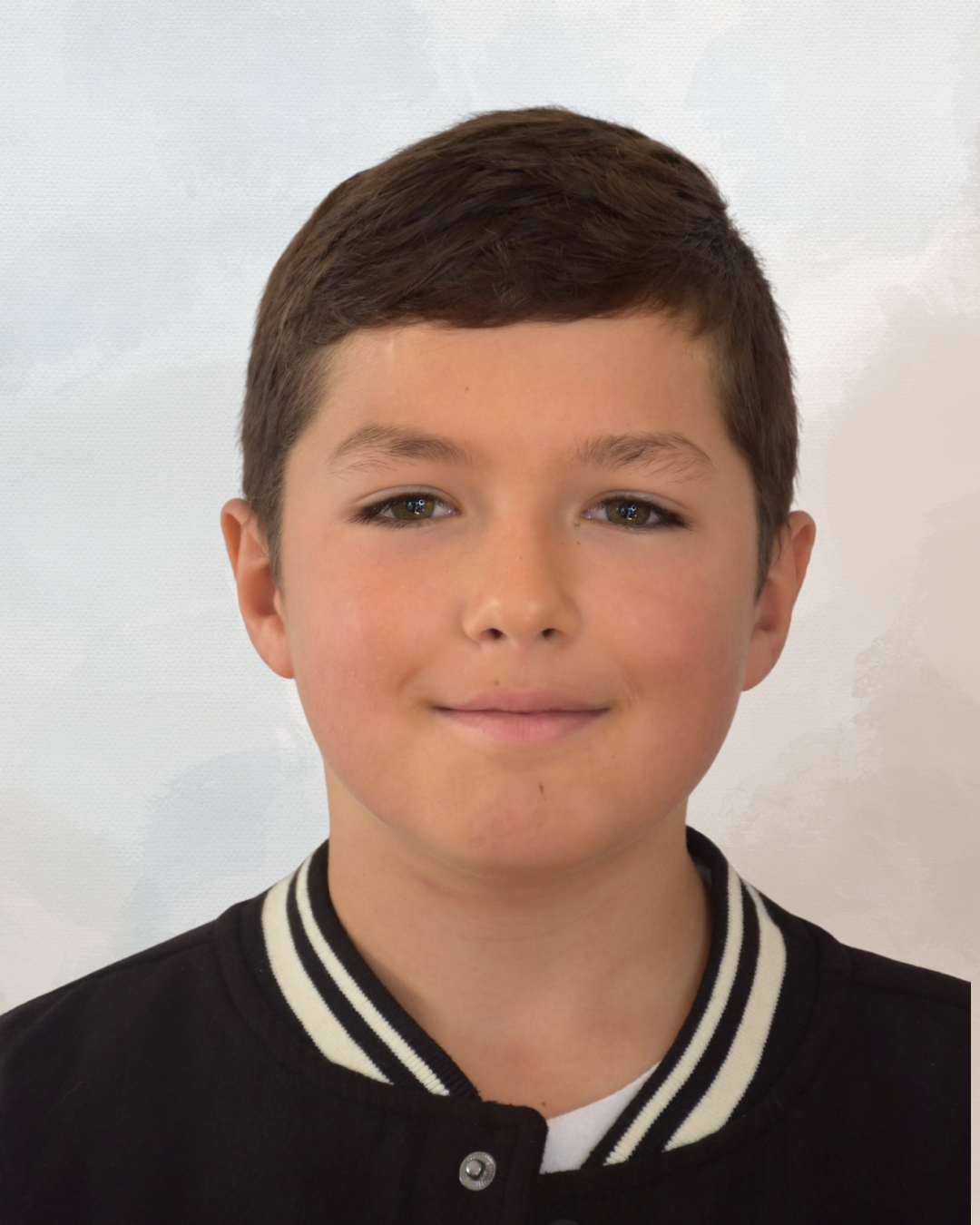 A young boy with brown hair and blue eyes, smiling softly, wearing a black and white varsity jacket, standing against a light background.