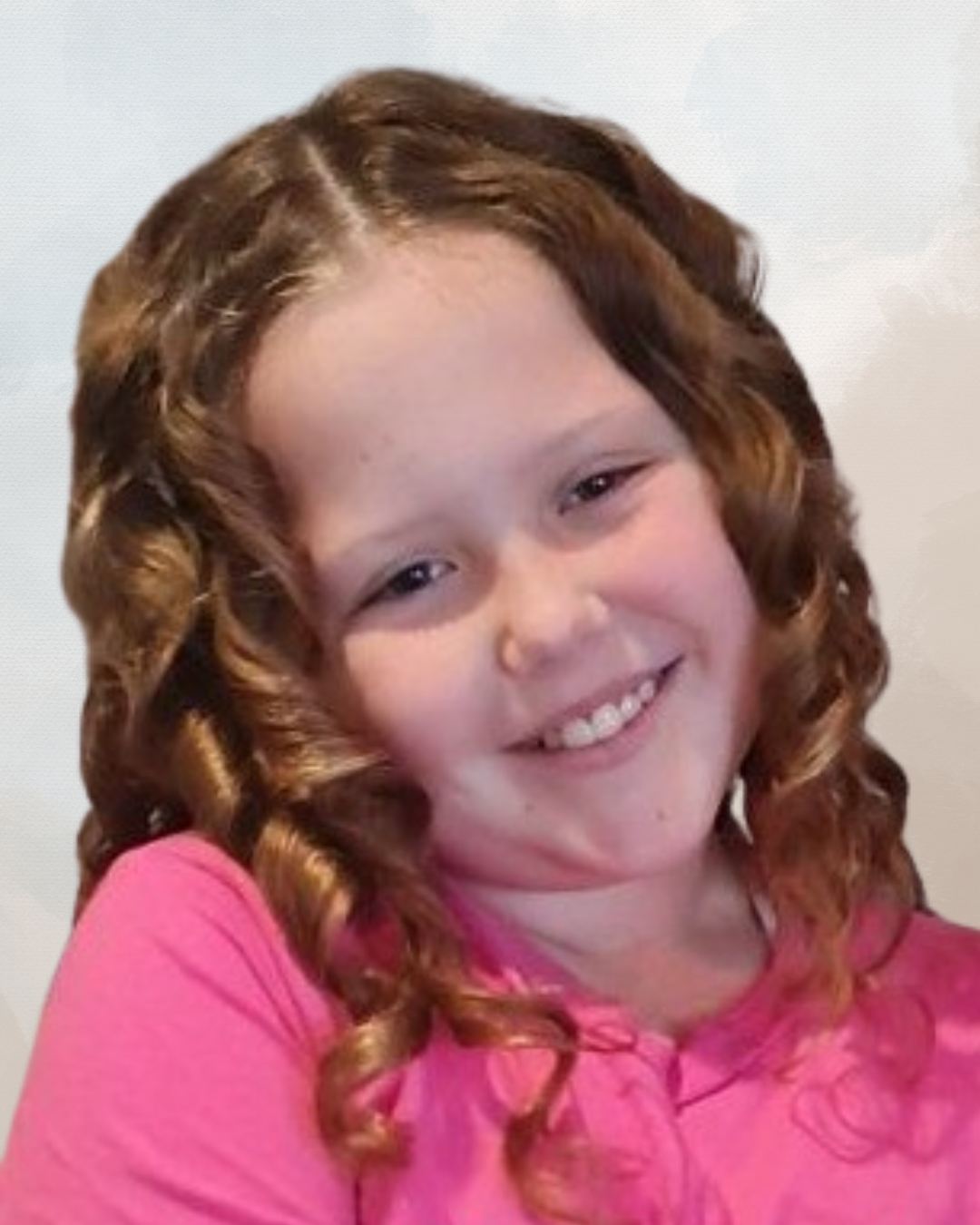 A young girl with red curly hair wearing a pink top, smiling.