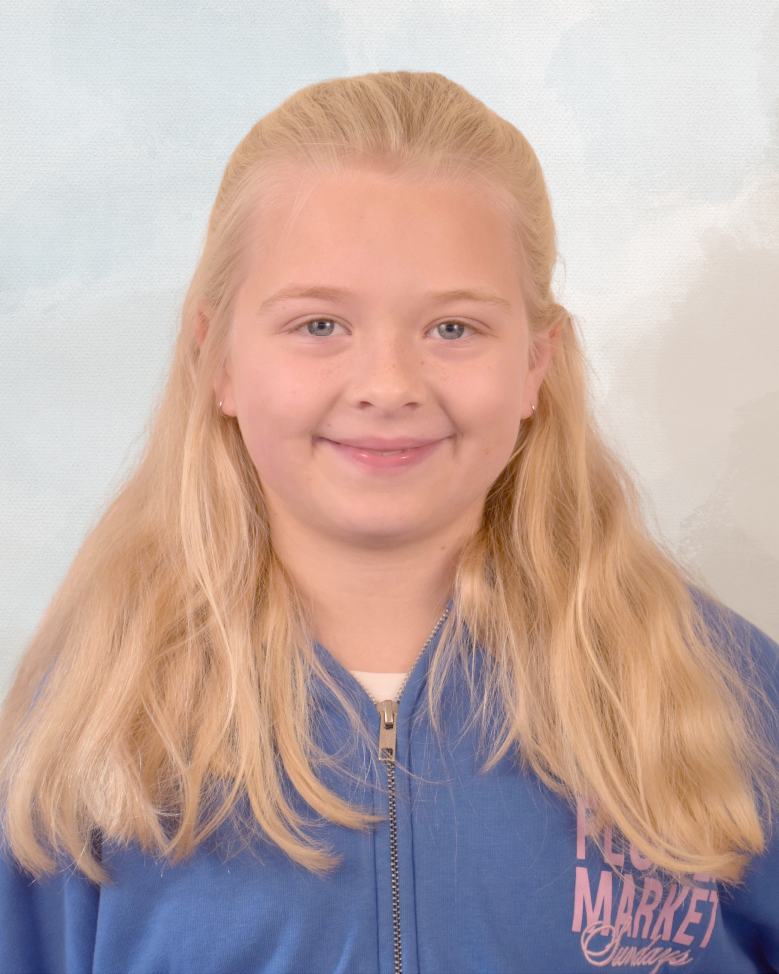A young girl with long blonde hair smiling, wearing a blue zip-up jacket with text on it, standing against a light background.