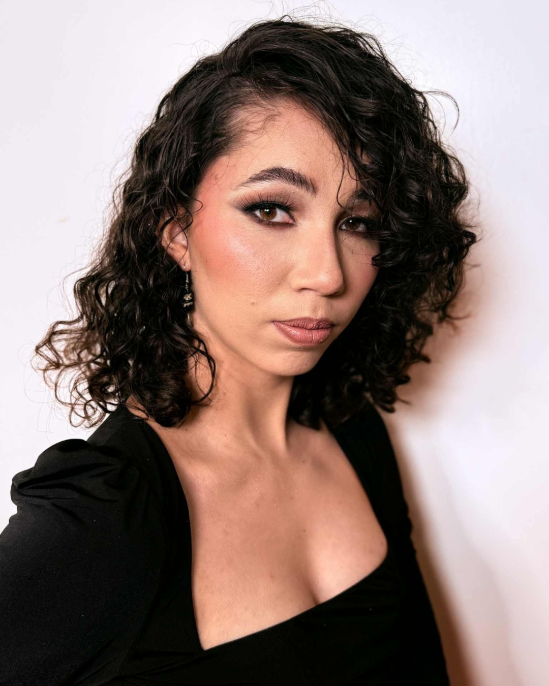 A woman with dark, curly hair and makeup making a confident expression.