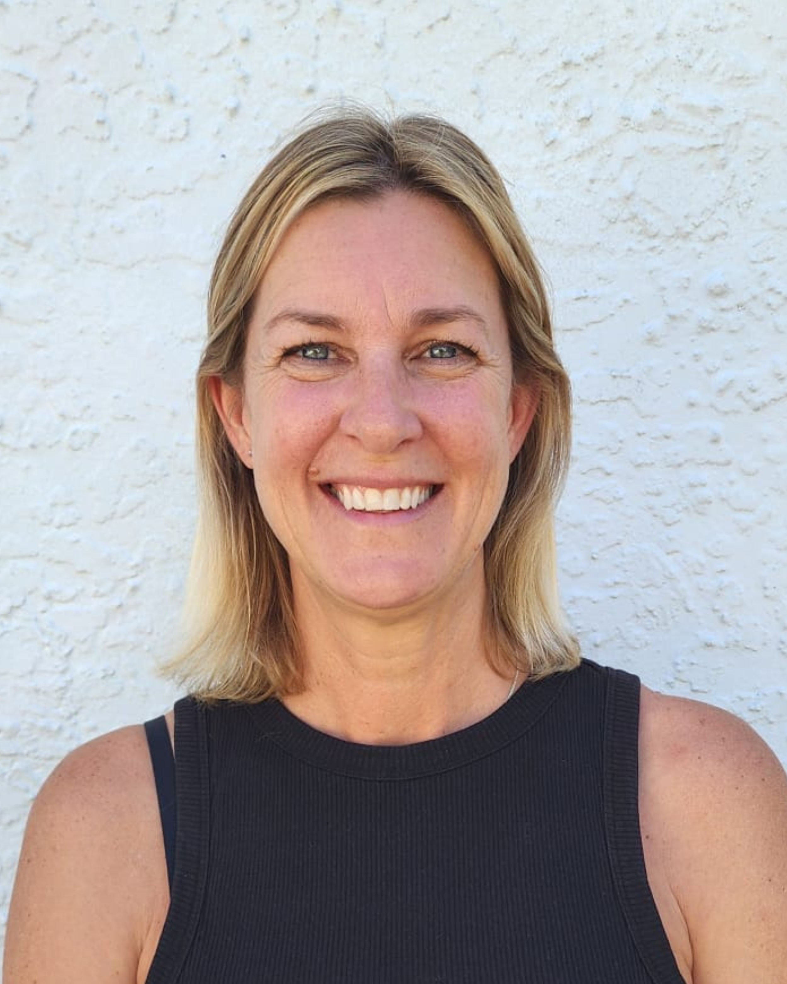 A smiling woman with shoulder-length blonde hair, wearing a sleeveless black top, standing against a white textured wall.