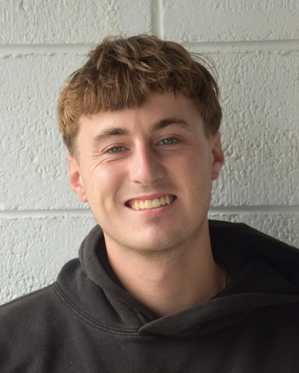 Young man with short brown hair smiling, wearing a black shirt, outdoors with cloudy sky background.