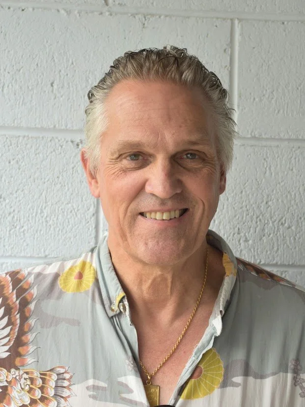 A middle-aged man with silver hair and blue eyes, smiling, wearing a patterned button-up shirt with yellow and brown accents, and a gold chain necklace, standing in front of a white brick wall.
