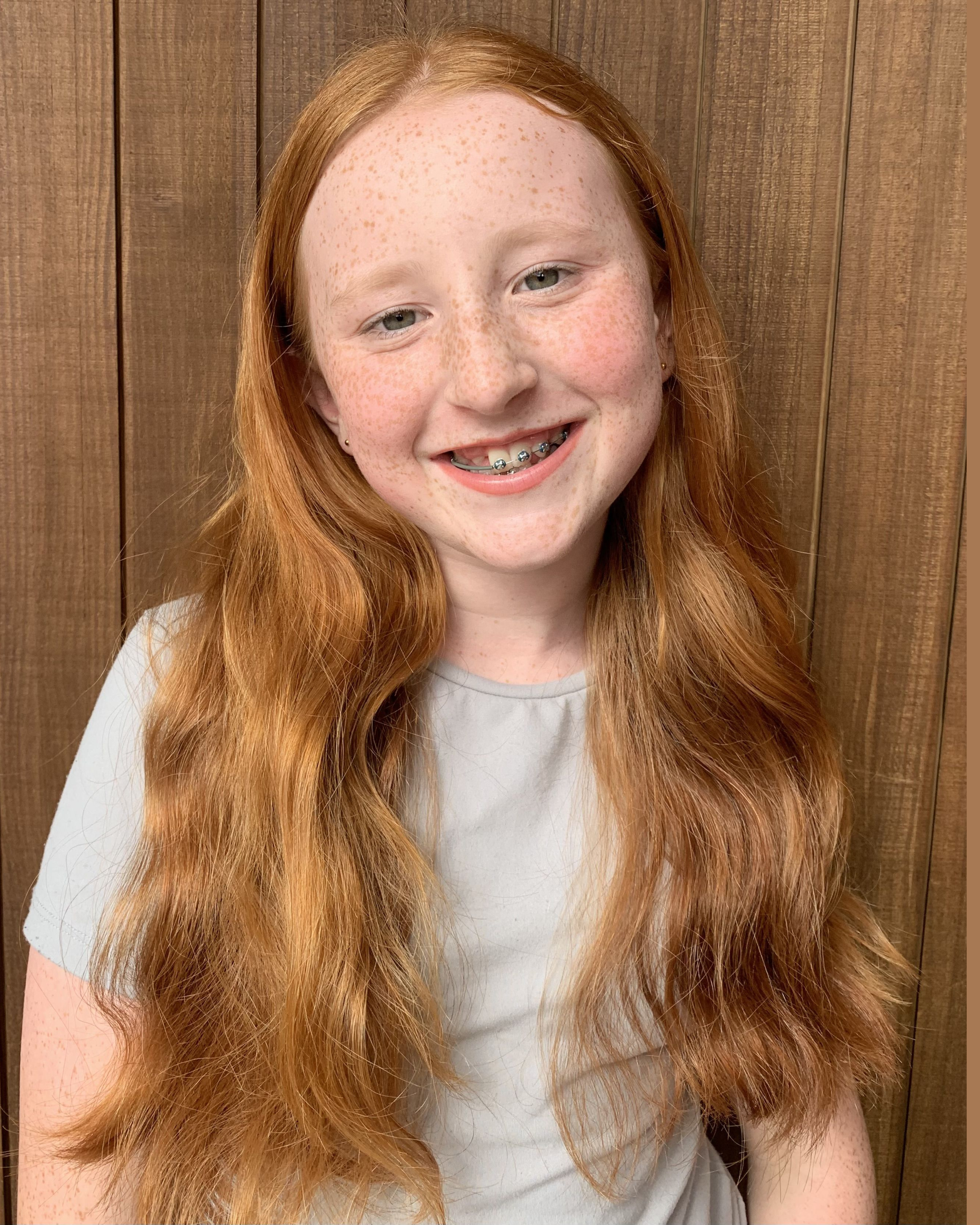 A young girl with long red hair and freckles smiling, showing braces on her teeth, wearing a light-colored shirt, standing in front of a wooden panel wall.