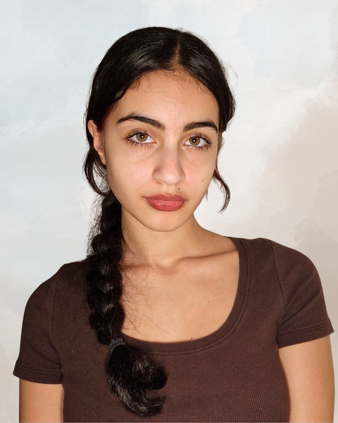 A young woman with long, dark hair in a braid, wearing a brown T-shirt, looking directly at the camera with a neutral expression.