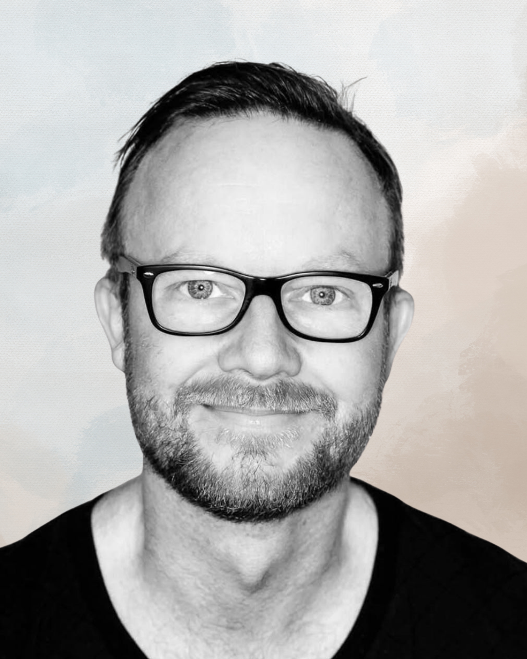 Black and white portrait of a man with glasses, short dark hair, a beard, and a smile, wearing a black shirt.