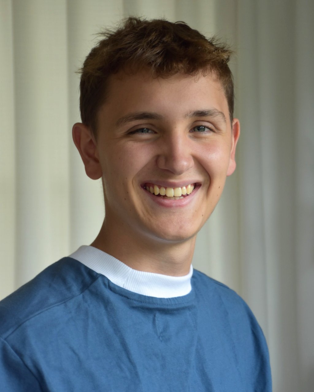 A young man with short dark hair and blue eyes standing outdoors in front of green trees, wearing a white sleeveless shirt with a black and white label that says 'ILABB'.