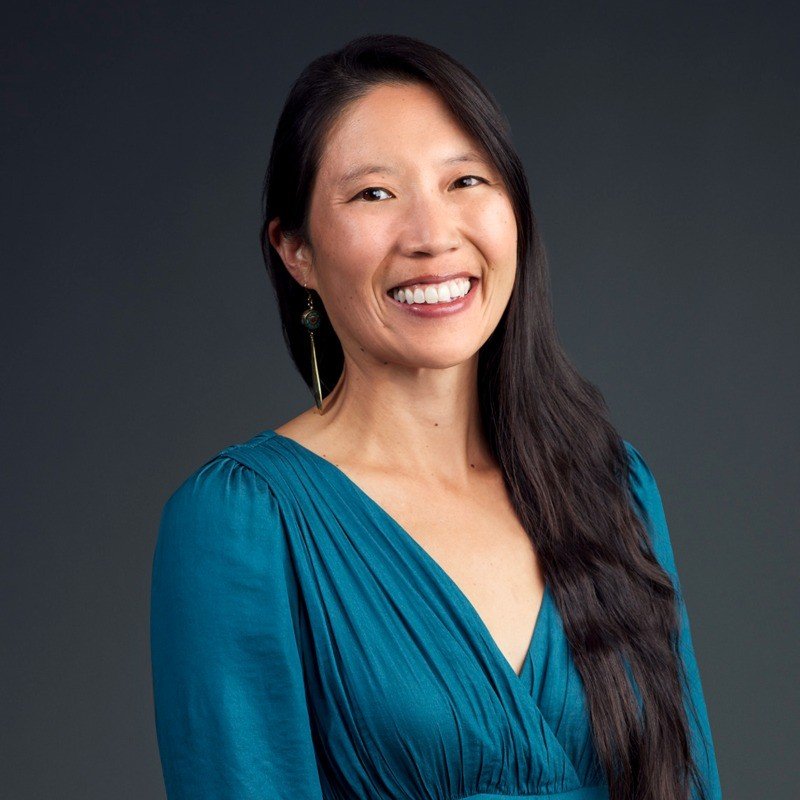 Smiling woman with long dark hair wearing a teal dress against a dark background