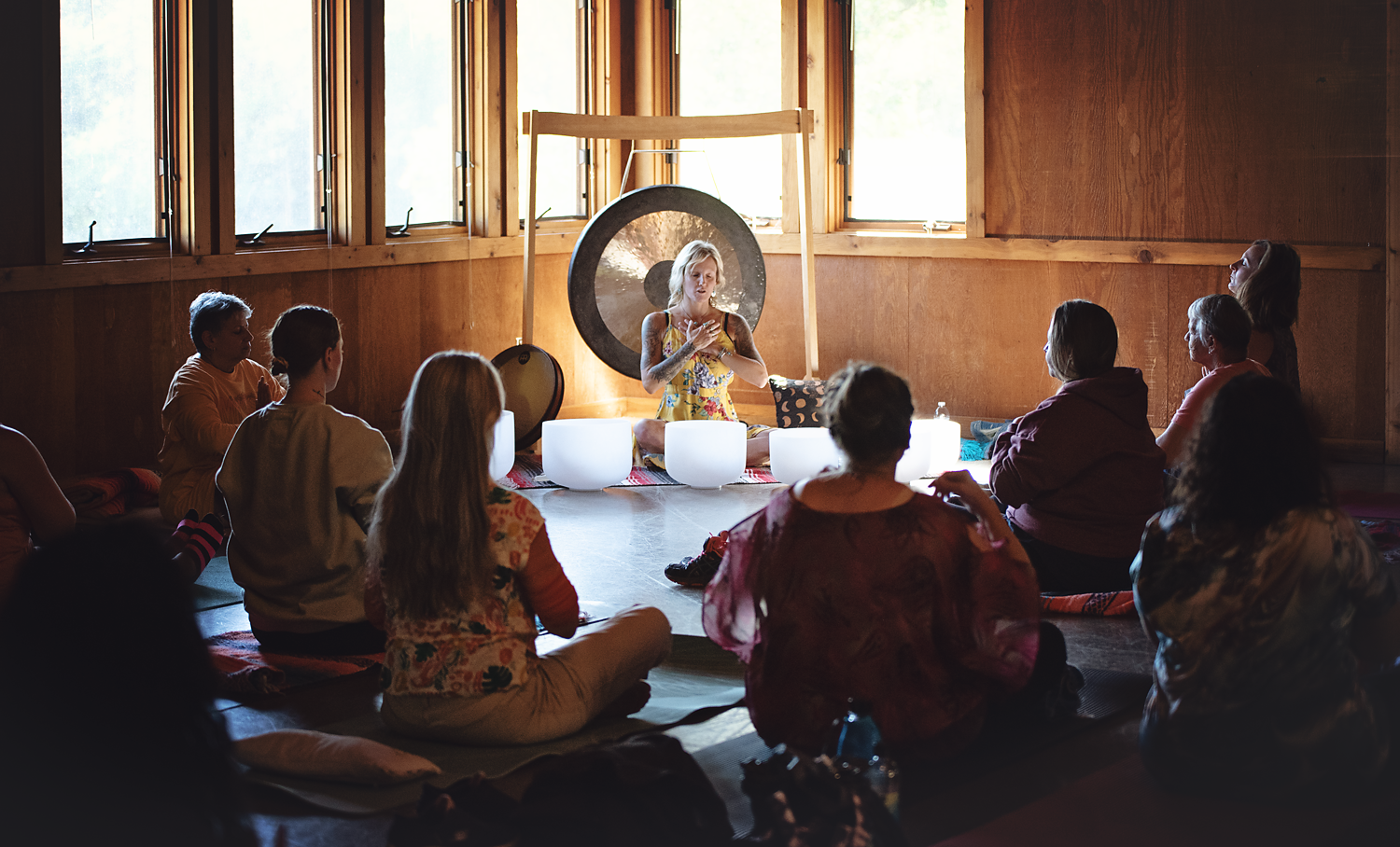 A woman leading a meditation or sound healing session with a gong, surrounded by seated participants in a wooden room near large windows.