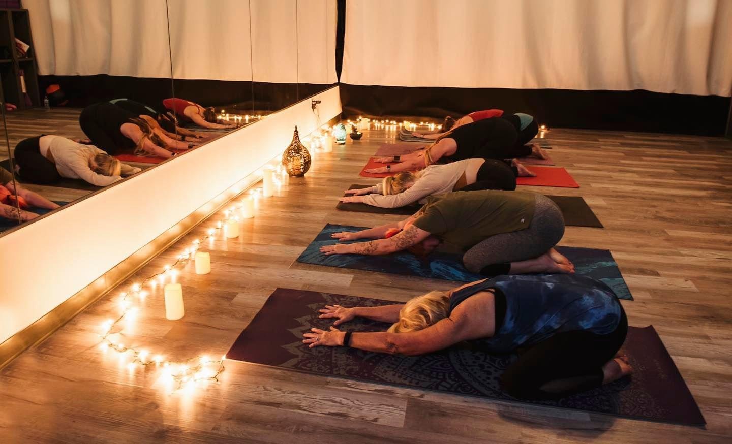 People participating in a yoga class, kneeling with their arms extended forward on yoga mats in a dimly lit room decorated with candles and drapes.