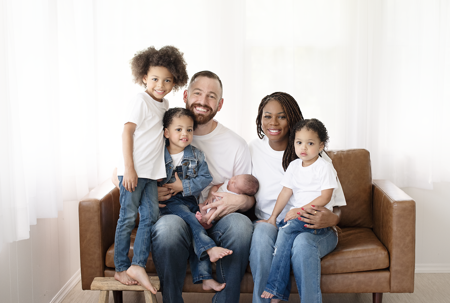 Multiracial family sitting on a brown couch in a bright room, holding a newborn baby, with three young girls around them.