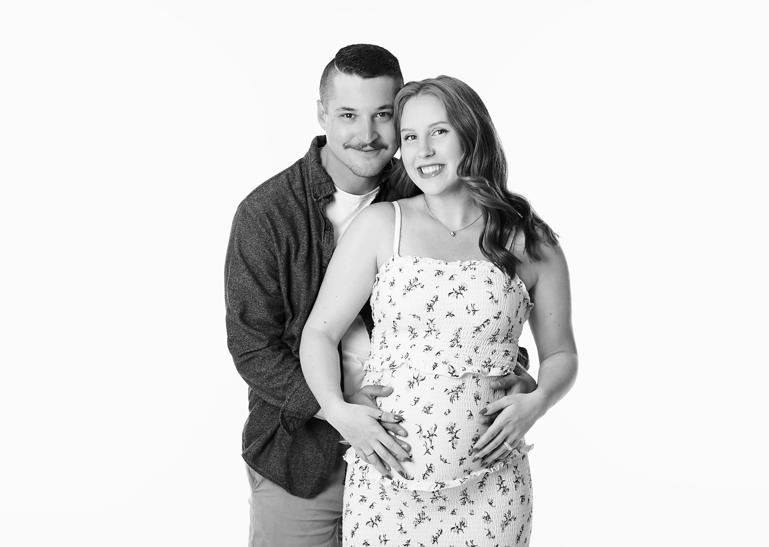 A black and white photo of a smiling couple. The man has short hair, a mustache, and is wearing a dark cardigan over a light shirt. The woman has long hair, and is wearing a sleeveless floral dress, with her hand resting on her pregnant belly, and bo