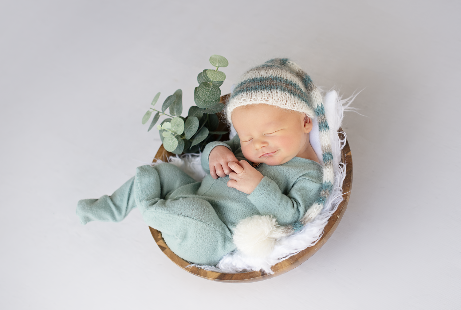 A sleeping baby in a green outfit and a knit hat, lying on a small wooden cradle with white bedding and eucalyptus leaves.