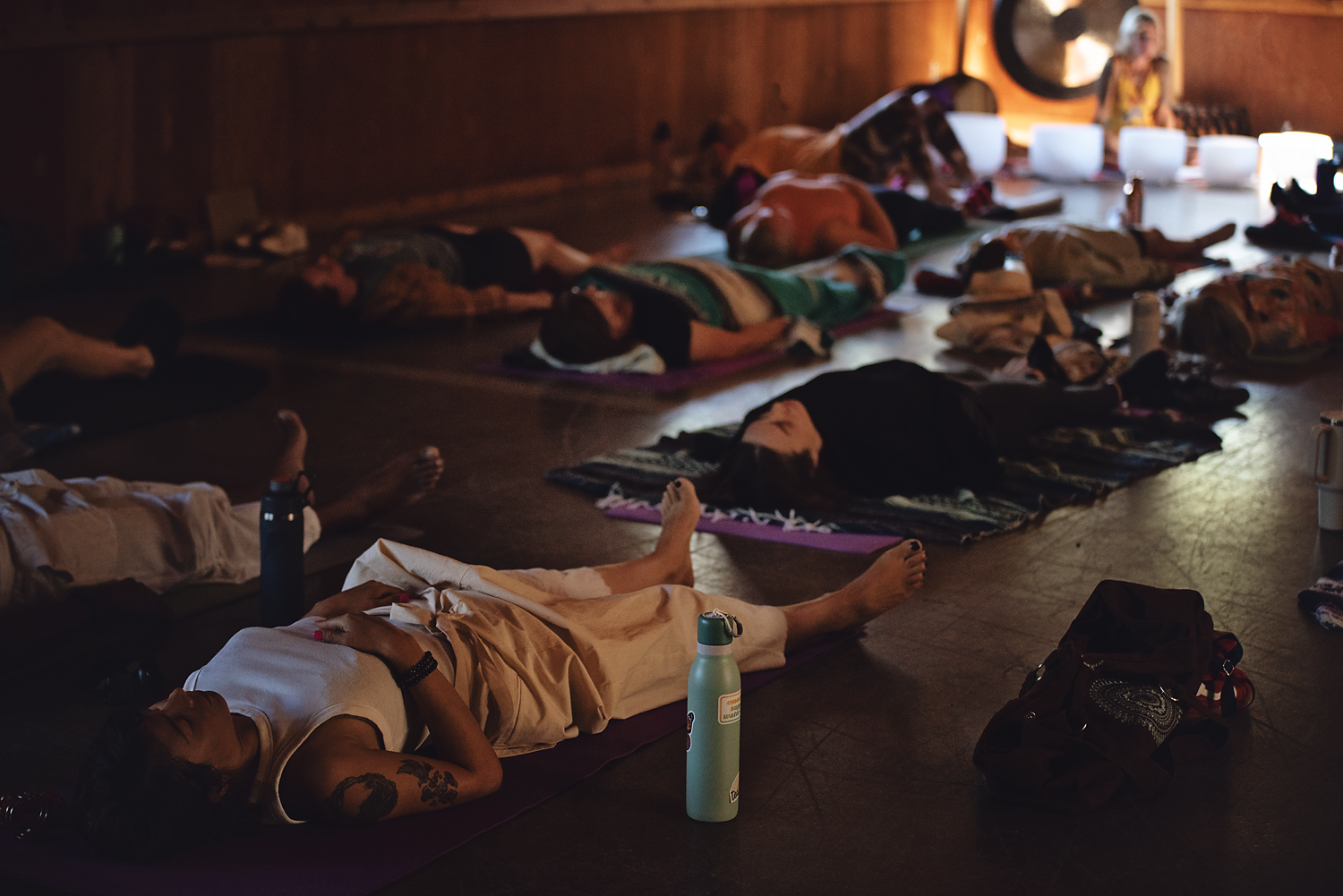 People lying down on yoga mats in a darkened indoor space, likely participating in a relaxation or meditation class.
