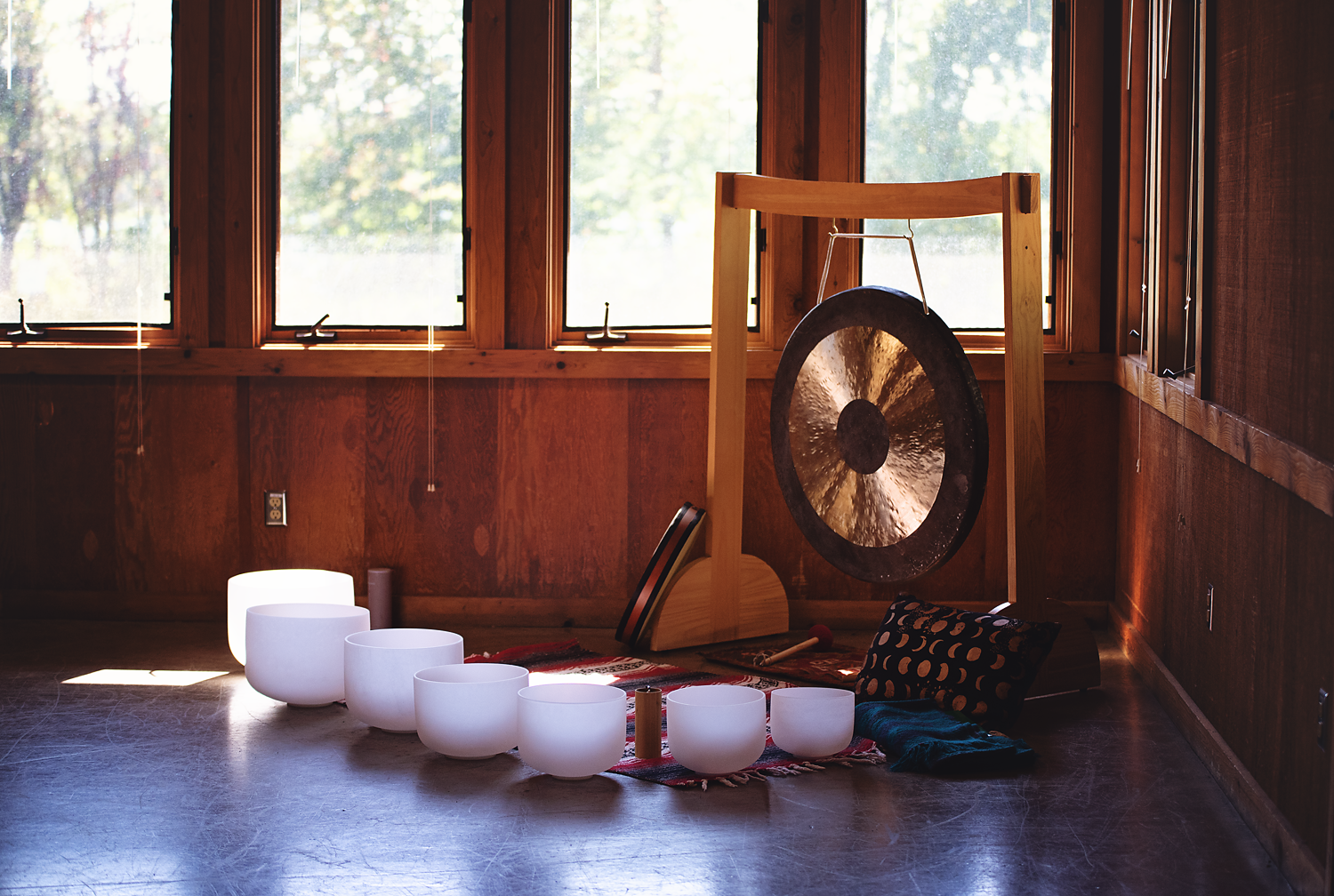 A collection of white singing bowls and a large gong in a wooden room with large windows showing trees outside, arranged on a rug with a cushion and drumsticks nearby.