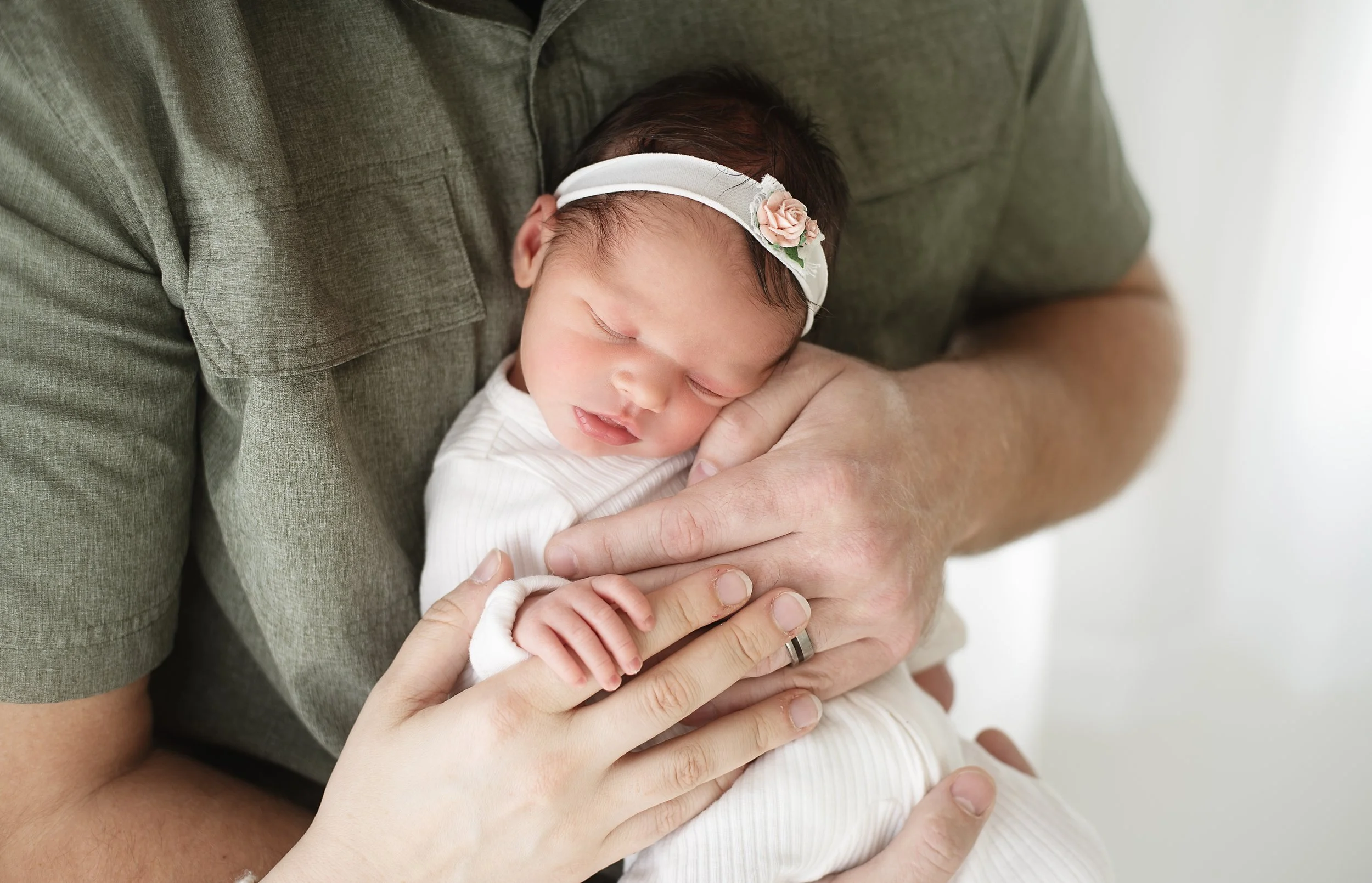 A baby girl sleeping peacefully on an adult's chest, wearing a white headband with a small pink flower, and dressed in a white outfit.
