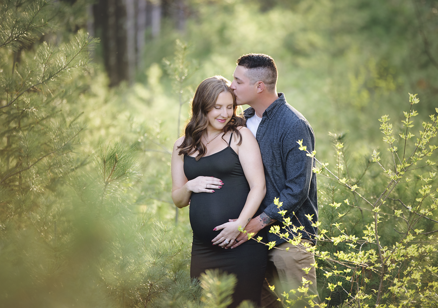 A pregnant woman in a black dress with a man in a blue shirt and beige pants standing in a green forest. The man is kissing her forehead, and she is smiling with closed eyes, holding her belly.