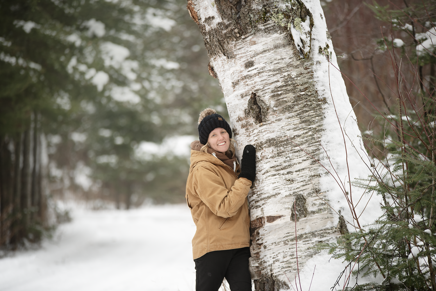 A woman wearing a brown jacket, black gloves, and a black knit hat holding and leaning against a large snow-covered birch tree in a snowy forest.