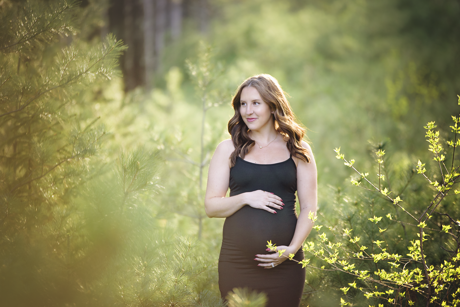 A pregnant woman with wavy brown hair wearing a black sleeveless dress, standing in a lush green forest with sunlight filtering through the trees, holding her belly with one hand and gently touching her stomach with the other.