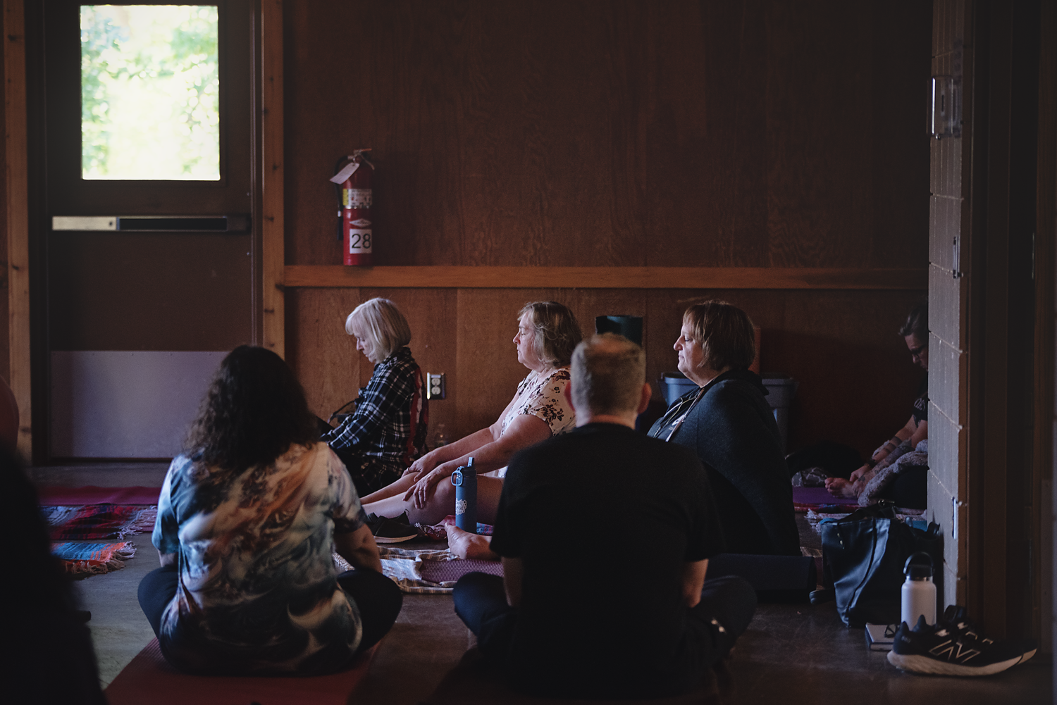 People sitting on the floor of a wooden room during a meditation or prayer session, facing the same direction with some holding their hands in their laps, with a fire extinguisher on the wall and a window letting in natural light.