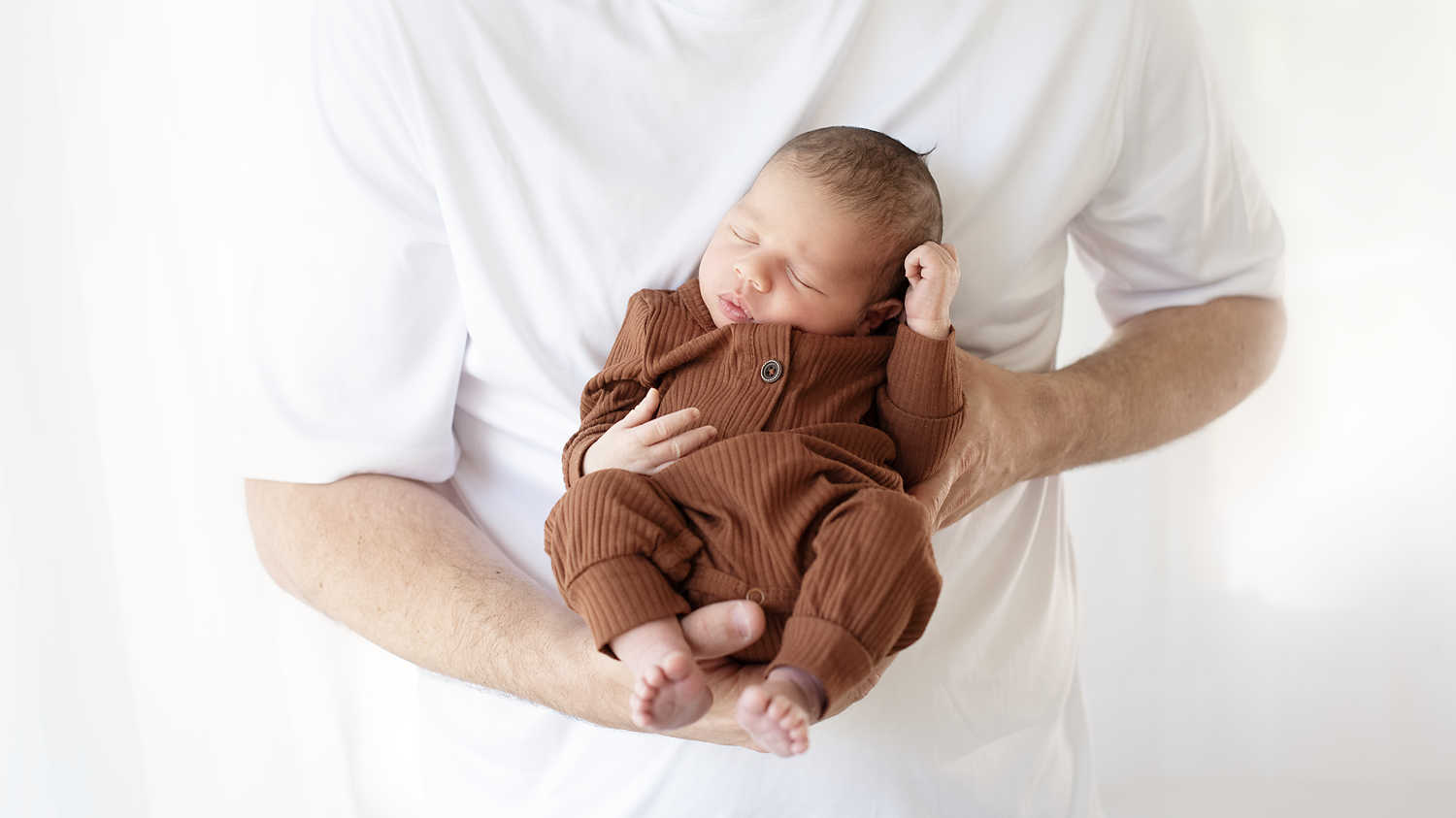 A baby sleeping peacefully in a person's arms, dressed in a brown outfit.
