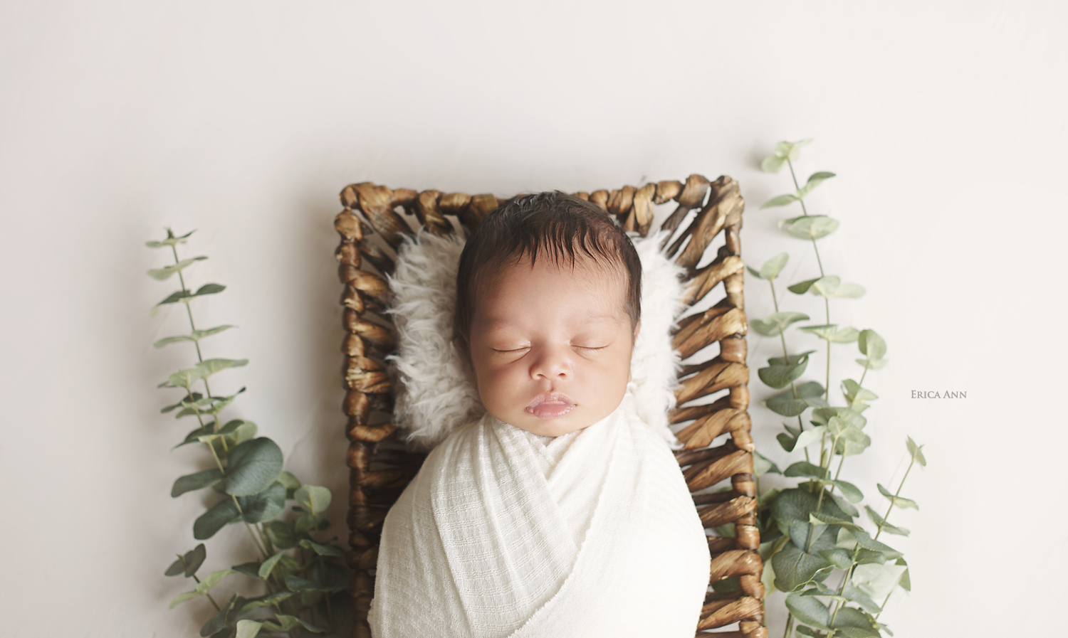 A sleeping newborn wrapped in a white blanket, lying on a furry pillow in a wicker basket with eucalyptus leaves on a white background.