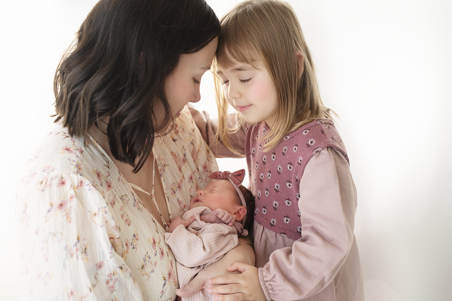 A woman holding a newborn baby, with a young girl leaning in close, all with their eyes closed in a tender moment.