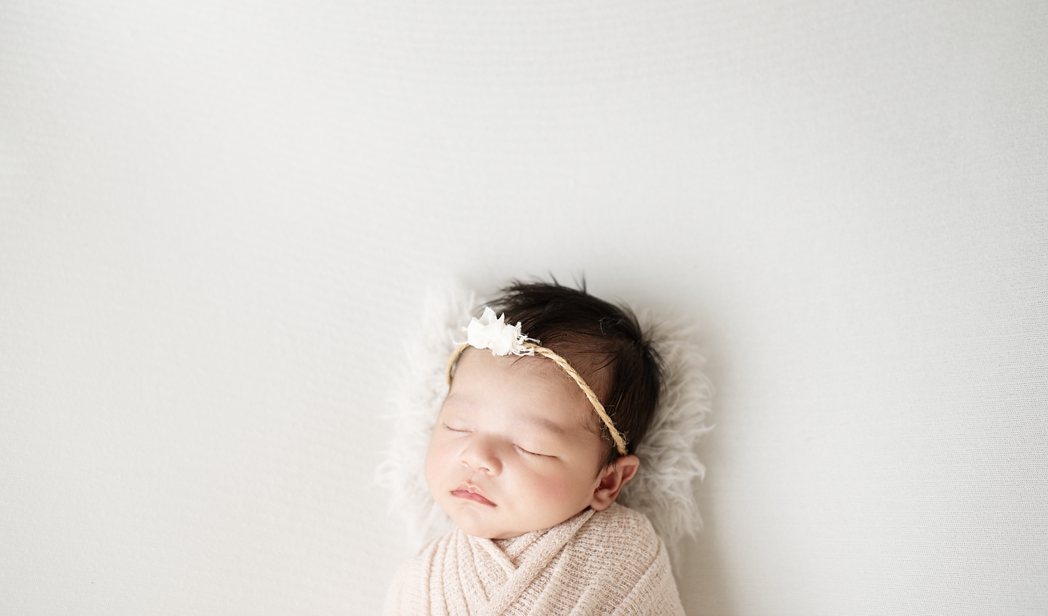 A sleeping newborn baby with dark hair, wrapped in a beige blanket, lying on a white furry surface, wearing a headband with a white flower.