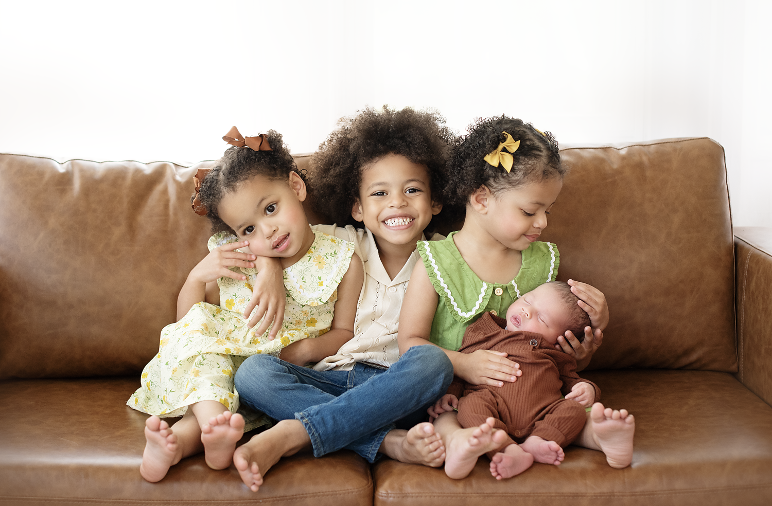 Four children sitting on a brown leather couch, with two girls and a boy smiling at the camera and a baby lying in the lap of the girl on the right, who holds the baby's head gently.
