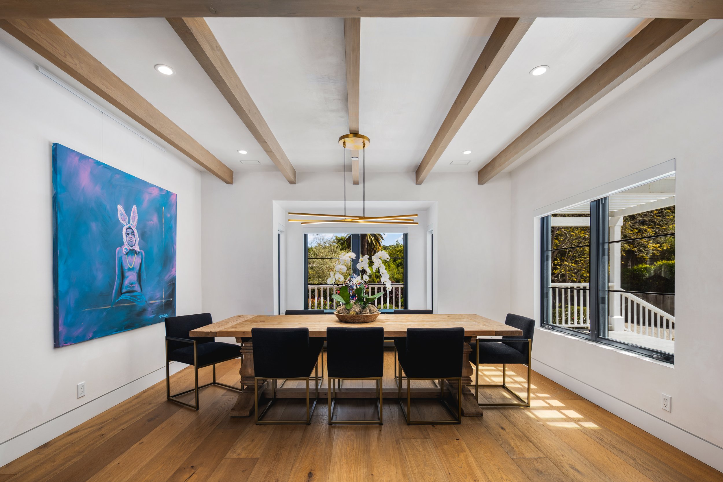 Modern dining room with a wooden table and chairs, a blue artwork on the wall, and large windows.