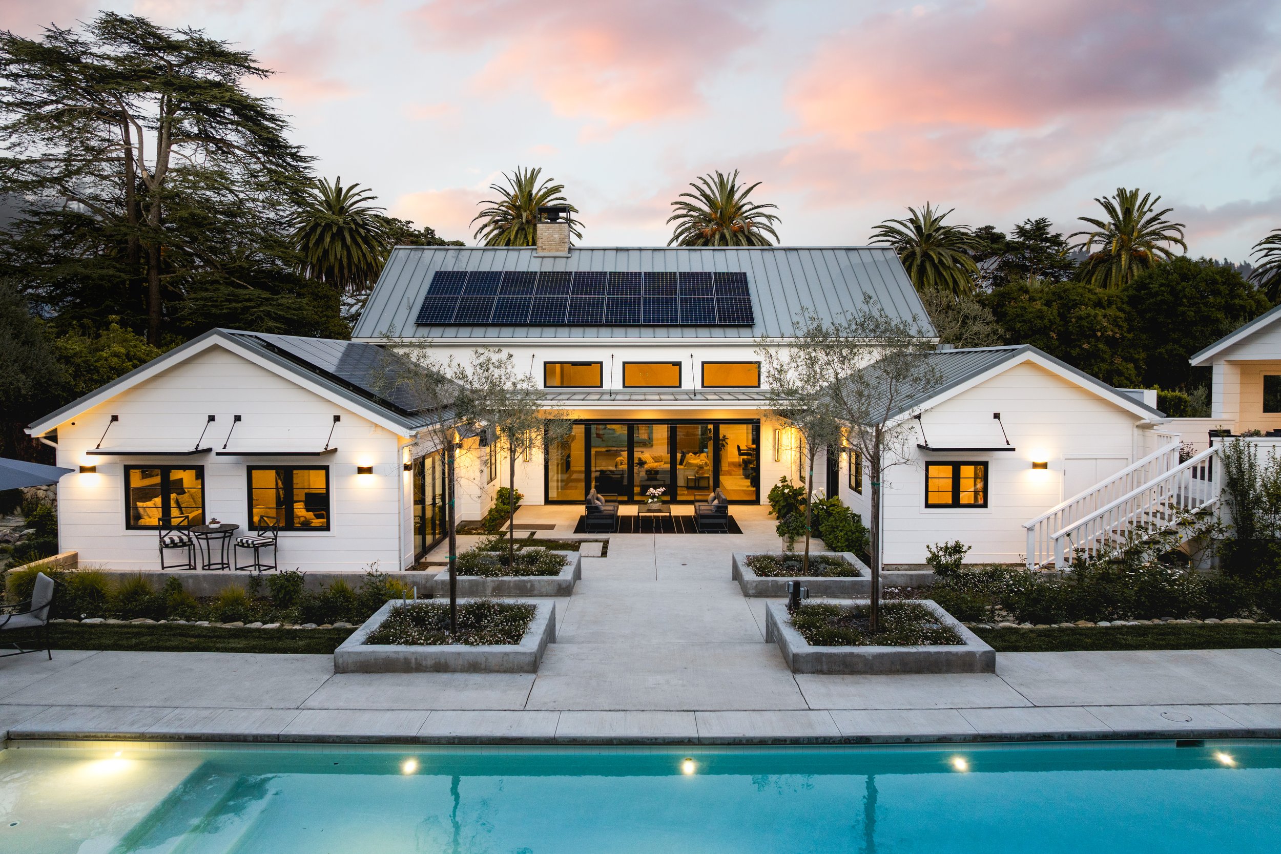 Modern house with solar panels on roof, surrounded by palm trees, a pool in the foreground, and lit-up interior visible through large windows.