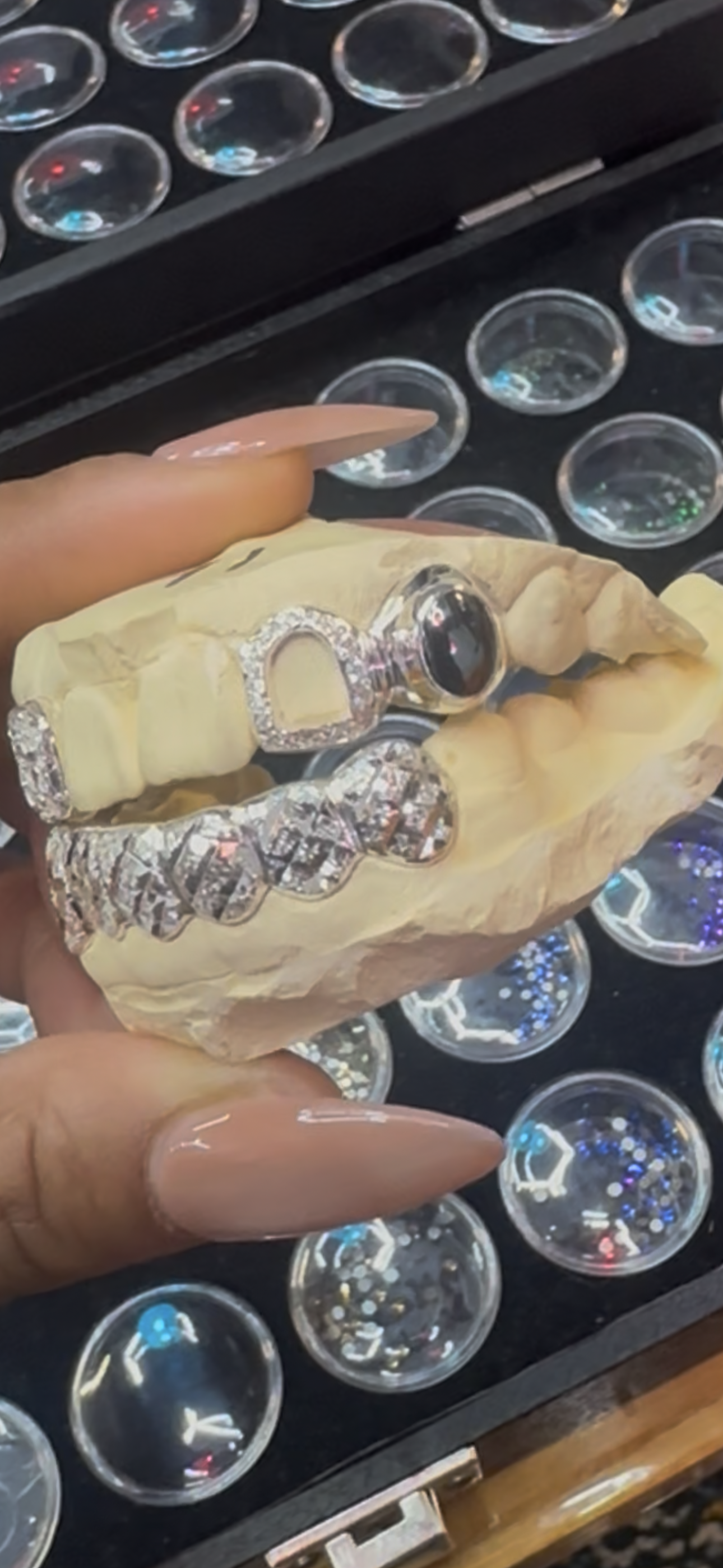 A jewelry display case with various round, colorful rhinestones or gems, and a hand holding a jewelry piece with a diamond-encrusted design and a black gemstone, placed on a concrete jaw model.