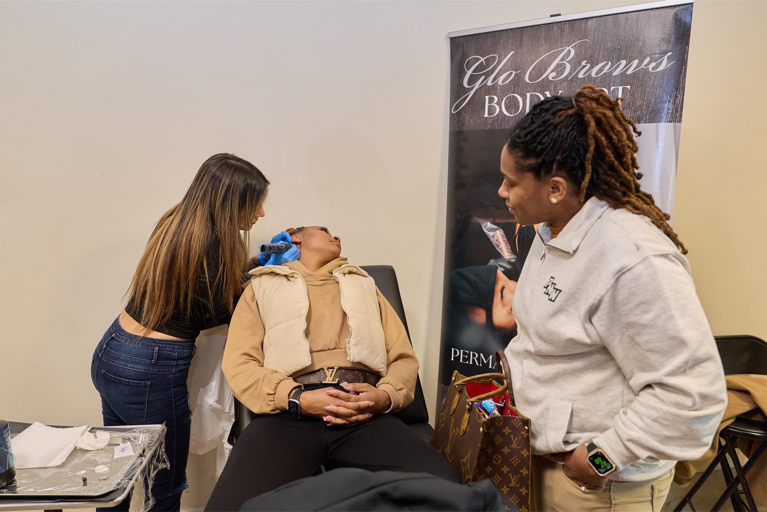 A woman lying back in a chair receiving a cosmetic procedure from a technician, with another woman observing nearby. A vertical banner with text stands in the background at at Jem Junkies Tooth Gems and  Custom Grillz in Chicago 