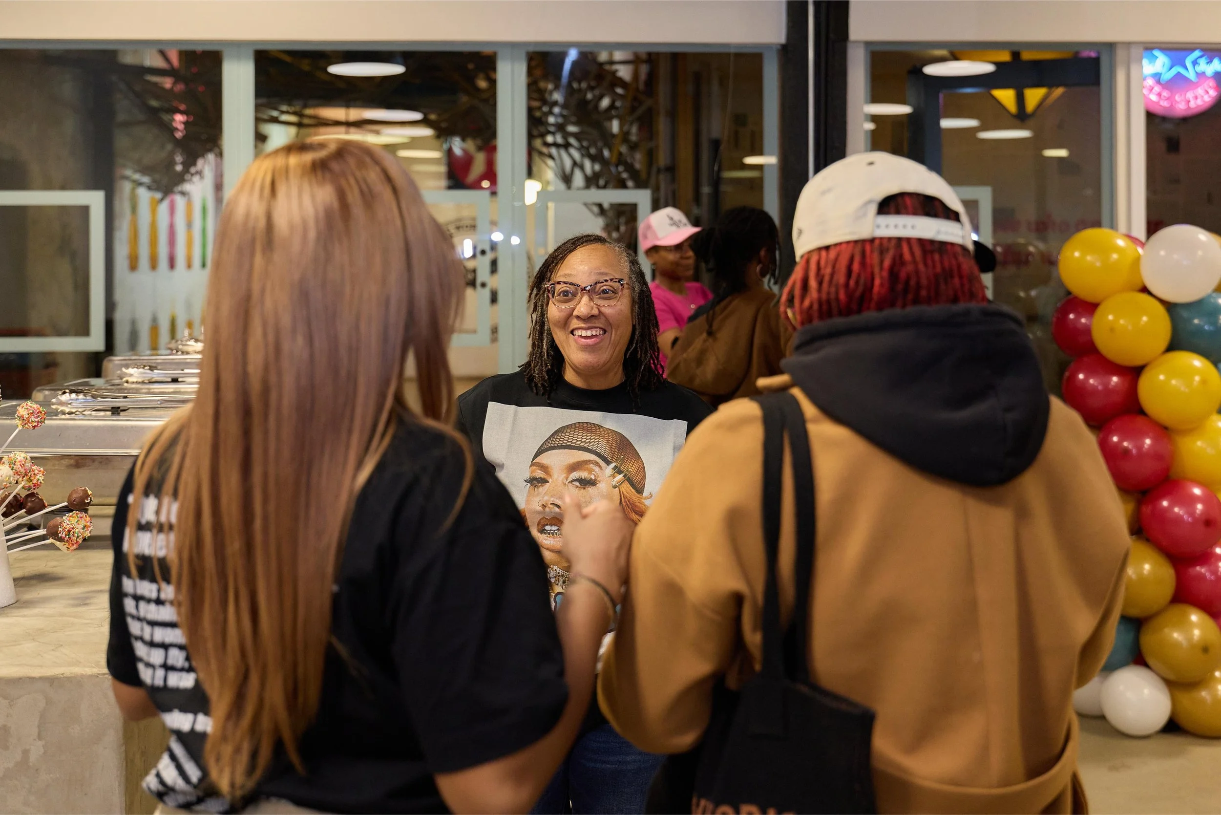 Three women saying hello and smiling at each other outside a bakery or cafe, with a woman in black with a graphic T-shirt in the center and two women with backpacks and caps, colorful balloons on the right, and desserts on the counter to the left.