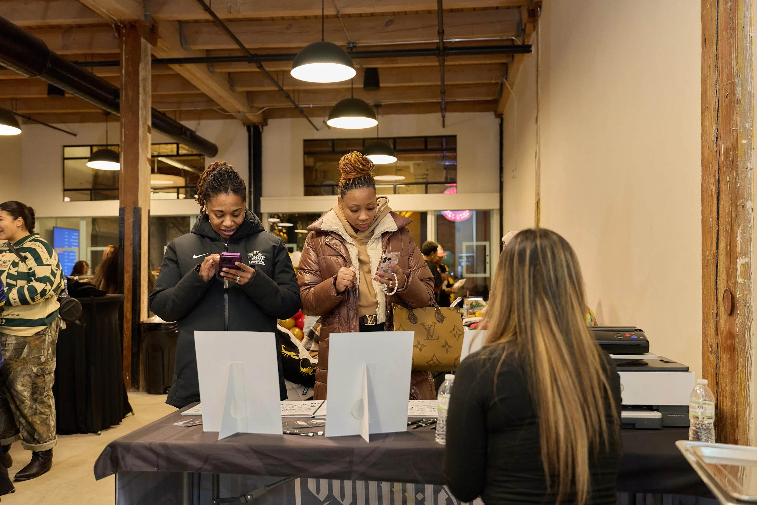 Two women standing in front of a table, looking at their phones, with a woman seated behind the table at Jem Junkies Tooth Gems and  Custom Grillz in Chicago 