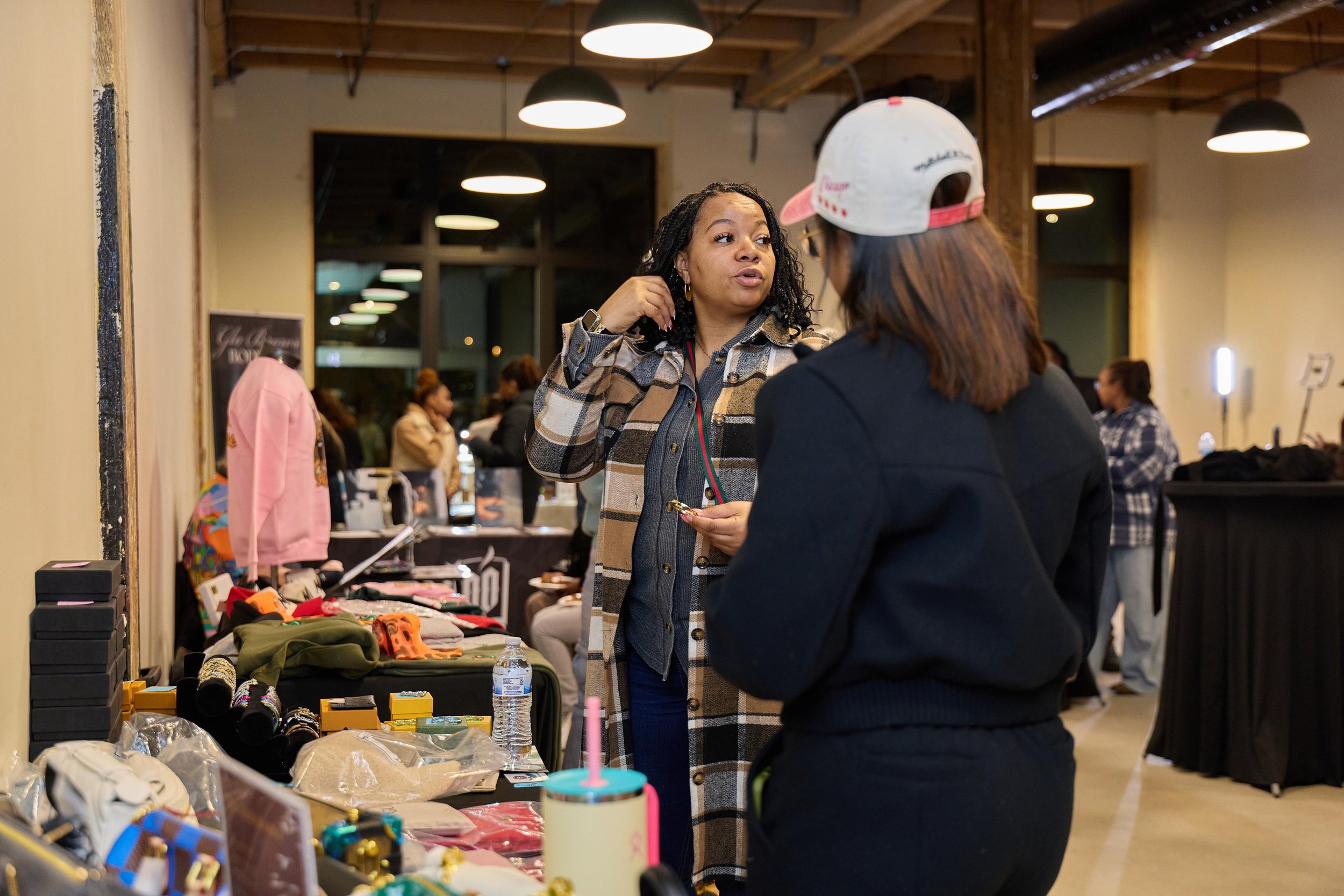 Two women having a conversation at a market stall with various items including clothing, jewelry, and a water bottle, inside a well-lit indoor space.
