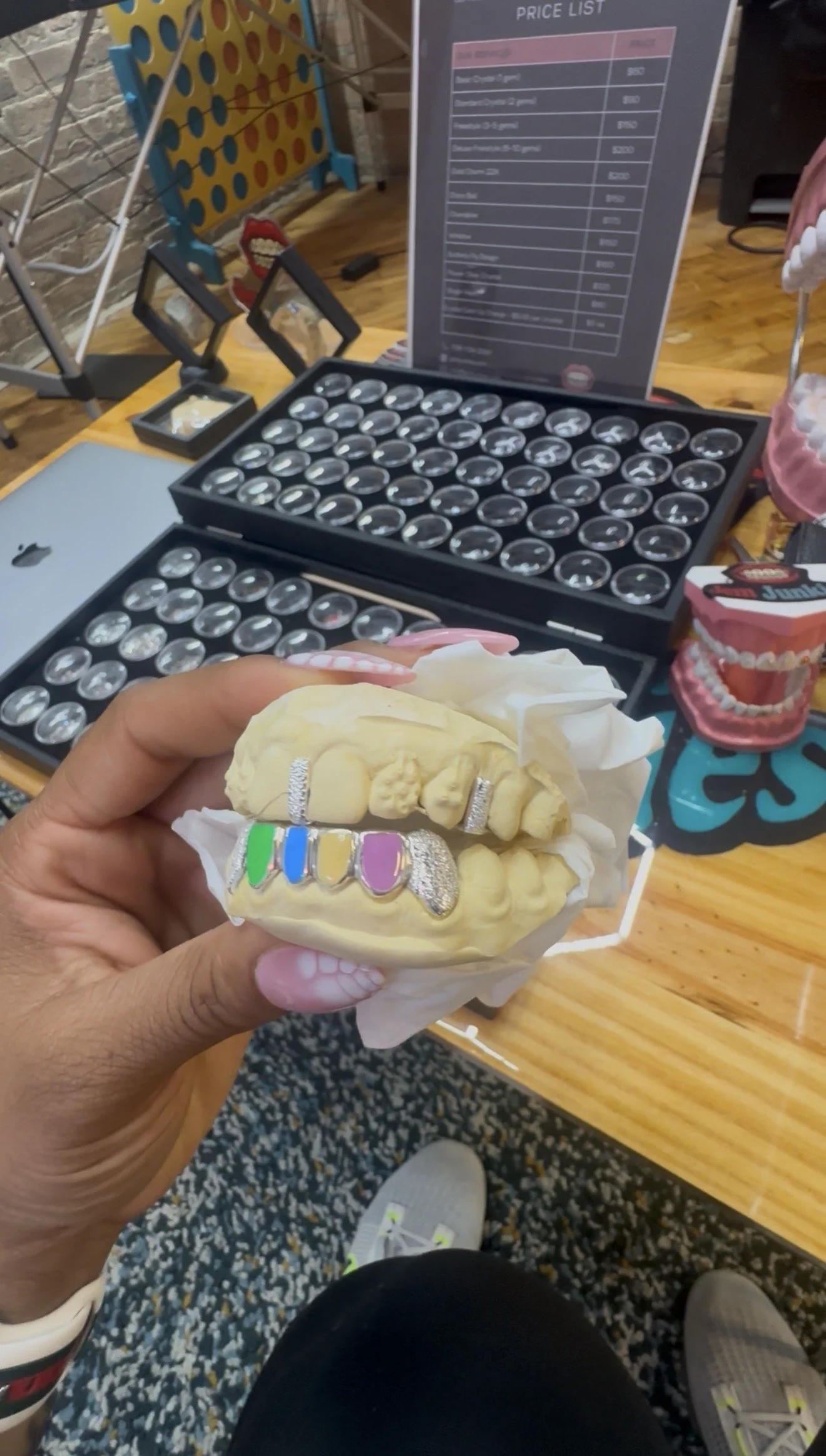 A person holding a dental mold with colorful textured teeth near a table with a price list and game pieces on it. The background includes a board game and a latte cup, and the setting appears to be an indoor café or game room.