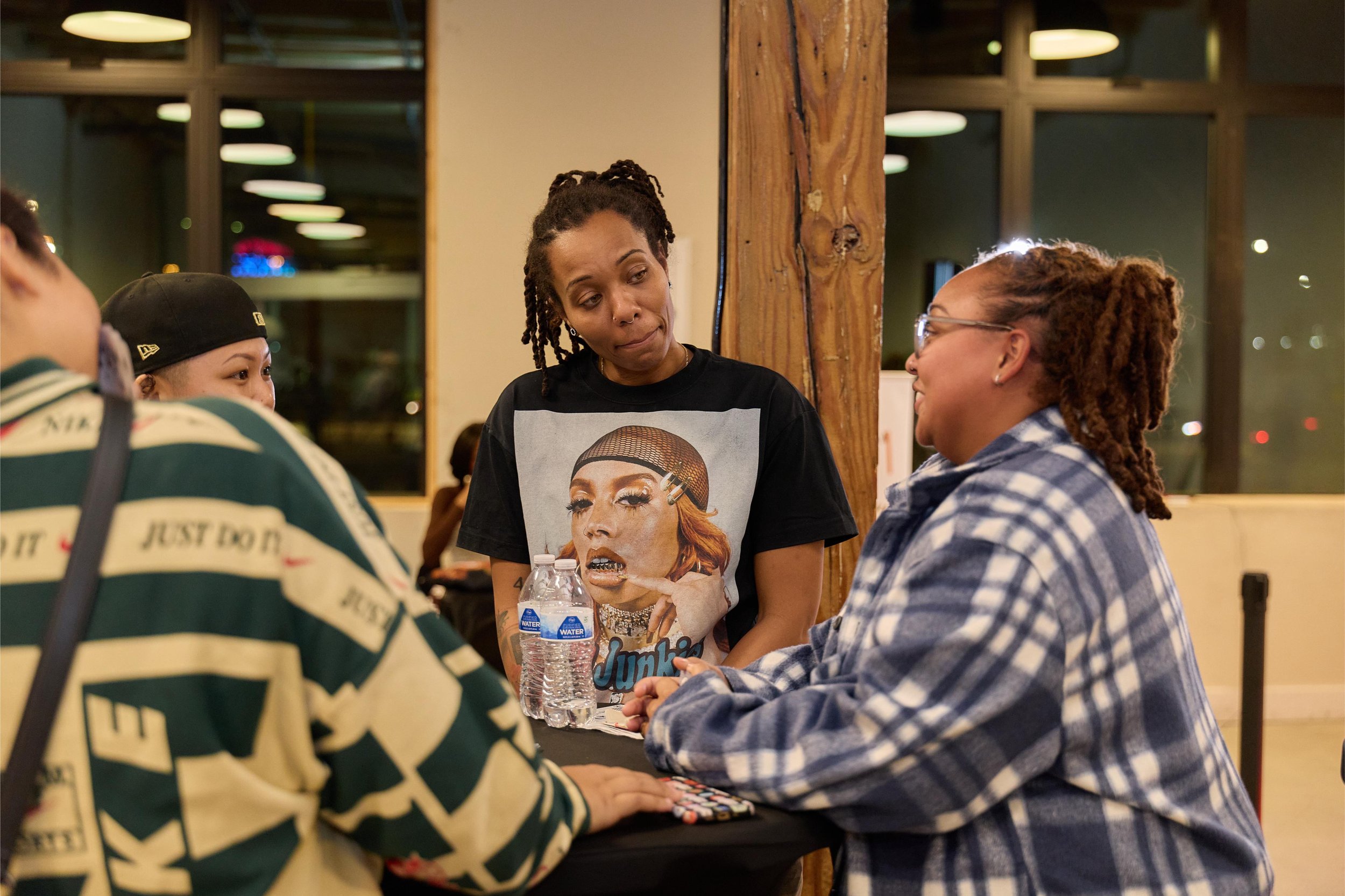 Three women engaged in conversation at a table inside a restaurant. One woman is wearing a graphic t-shirt and has dreadlocks, another is wearing a plaid shirt, and the third woman has a black beanieat Jem Junkies Tooth Gems and  Custom Grillz in Chi