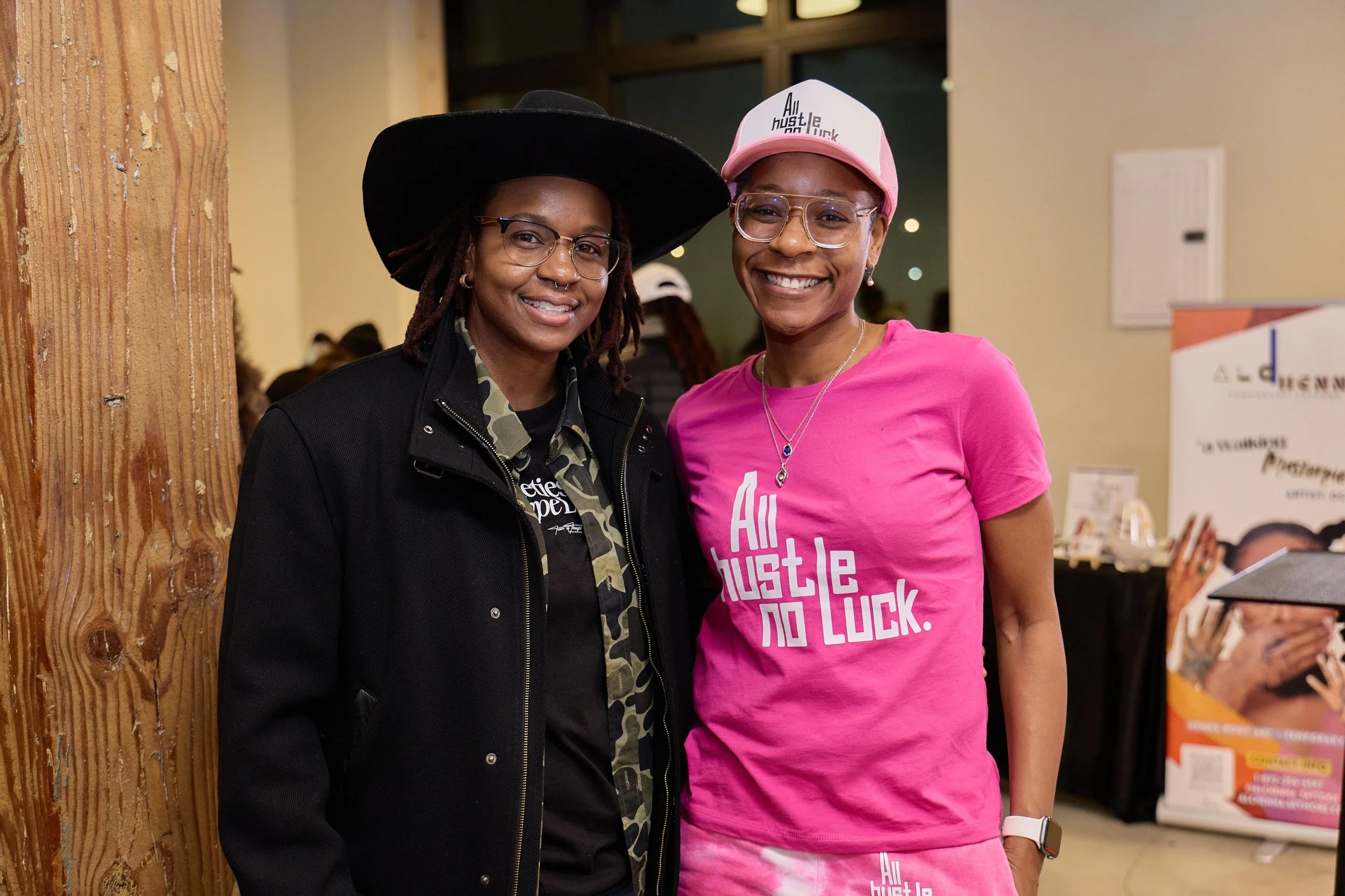 Two women smiling and posing together at an indoor event. One woman is wearing a black hat and black jacket with a camouflage shirt underneath. The other woman is wearing glasses, a pink cap, and a pink T-shirt that says "All Hustle No Luck." They ar